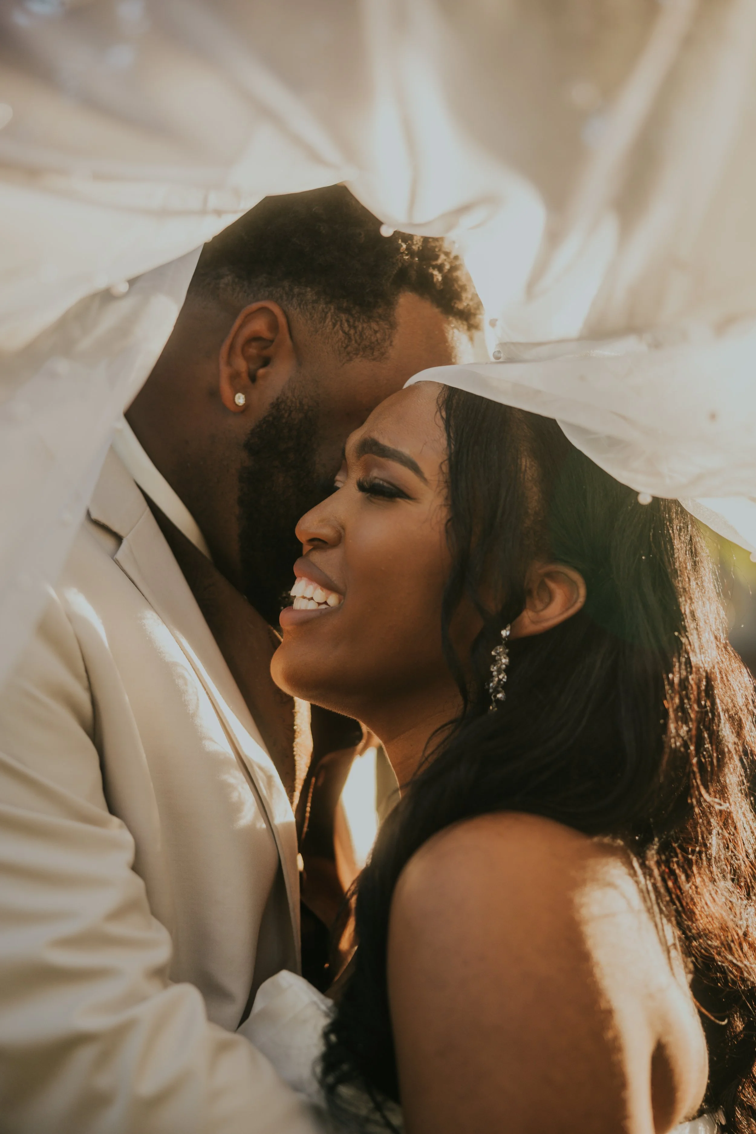A happy couple embracing outdoors, with sunlight illuminating their faces, under a white cloth canopy.