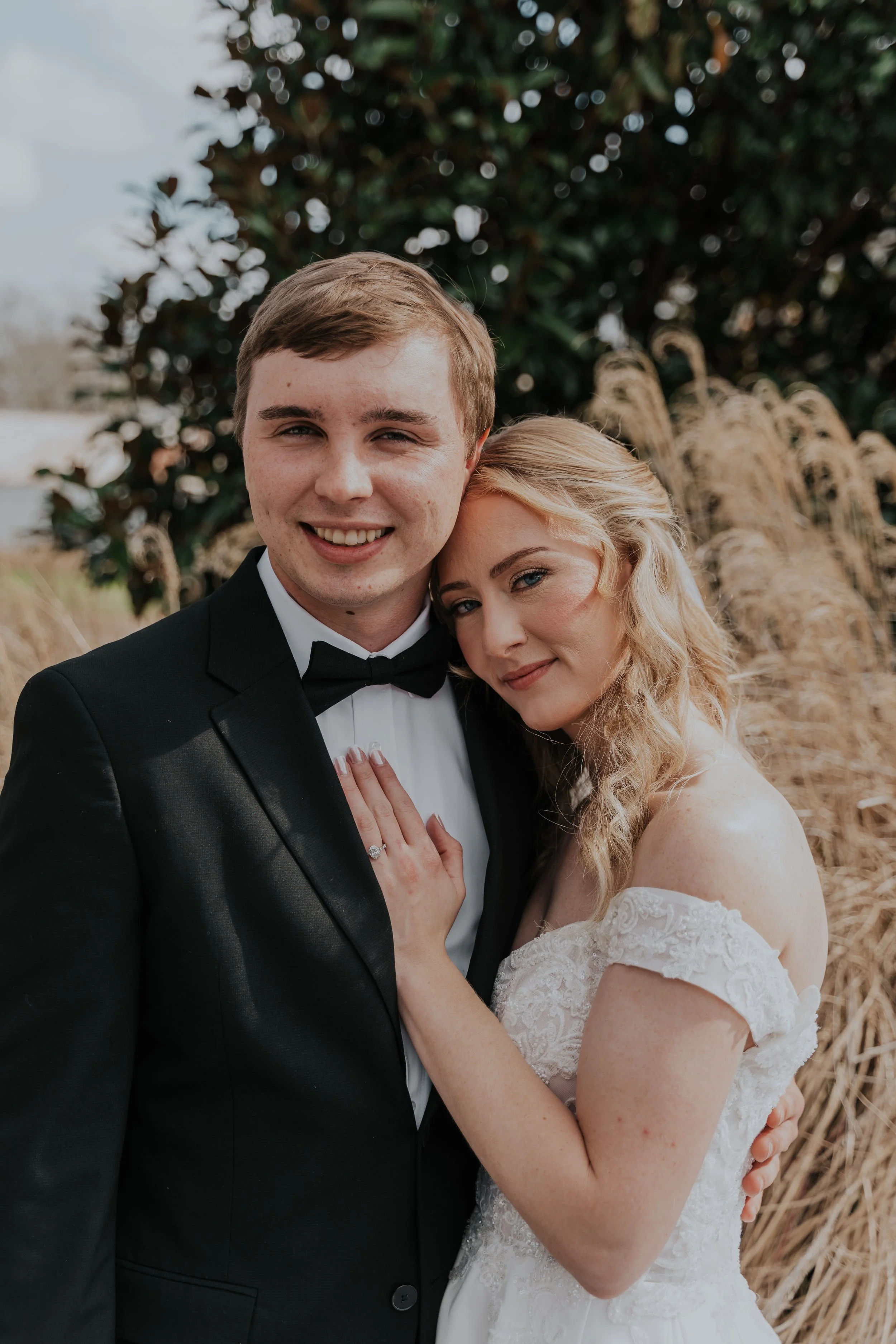 A newlywed couple in wedding attire, standing outdoors with a bush and tall grass background.