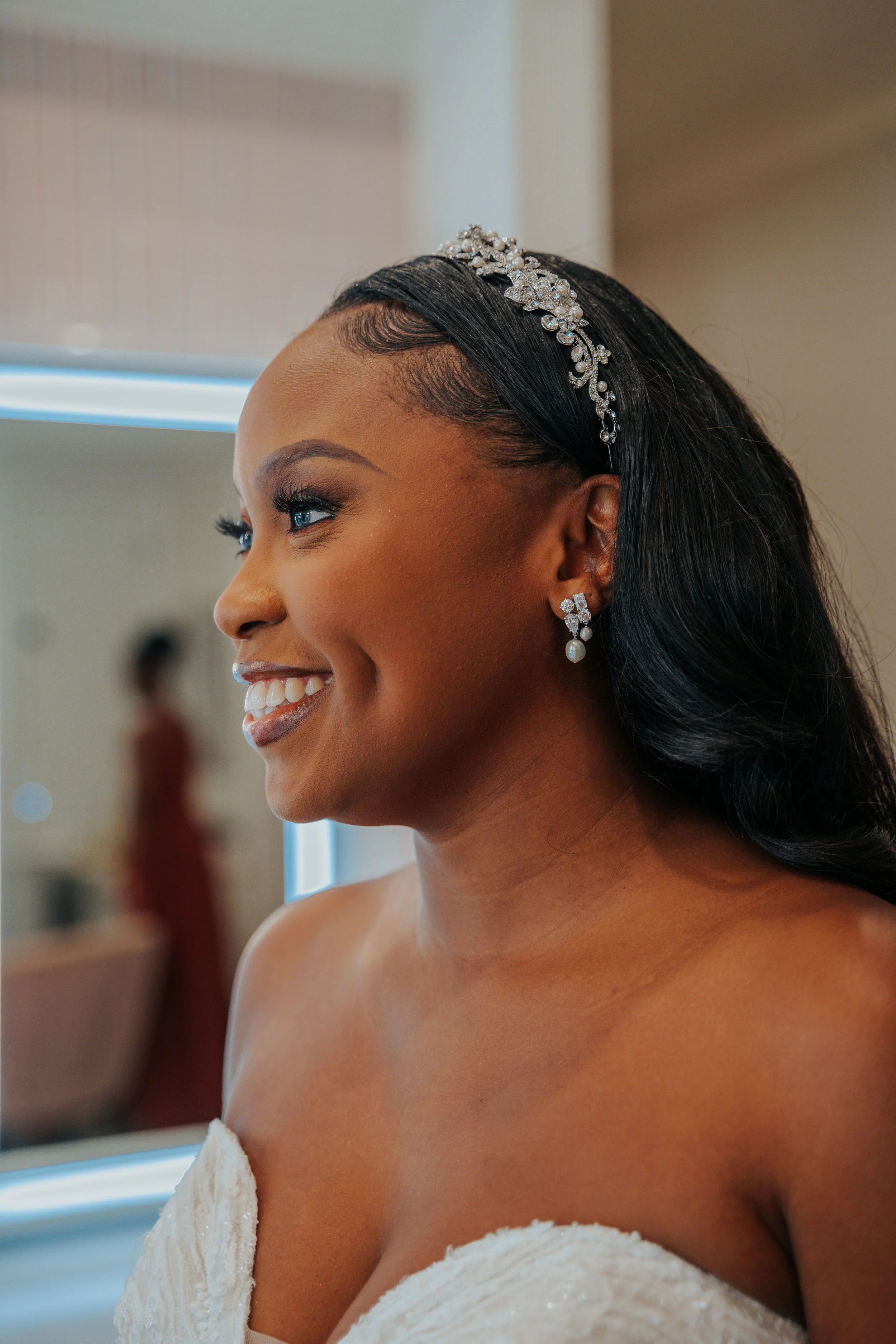Close-up of a woman with dark hair, wearing a tiara and pearl earrings, smiling in front of a mirror, dressed in a strapless white gown.
