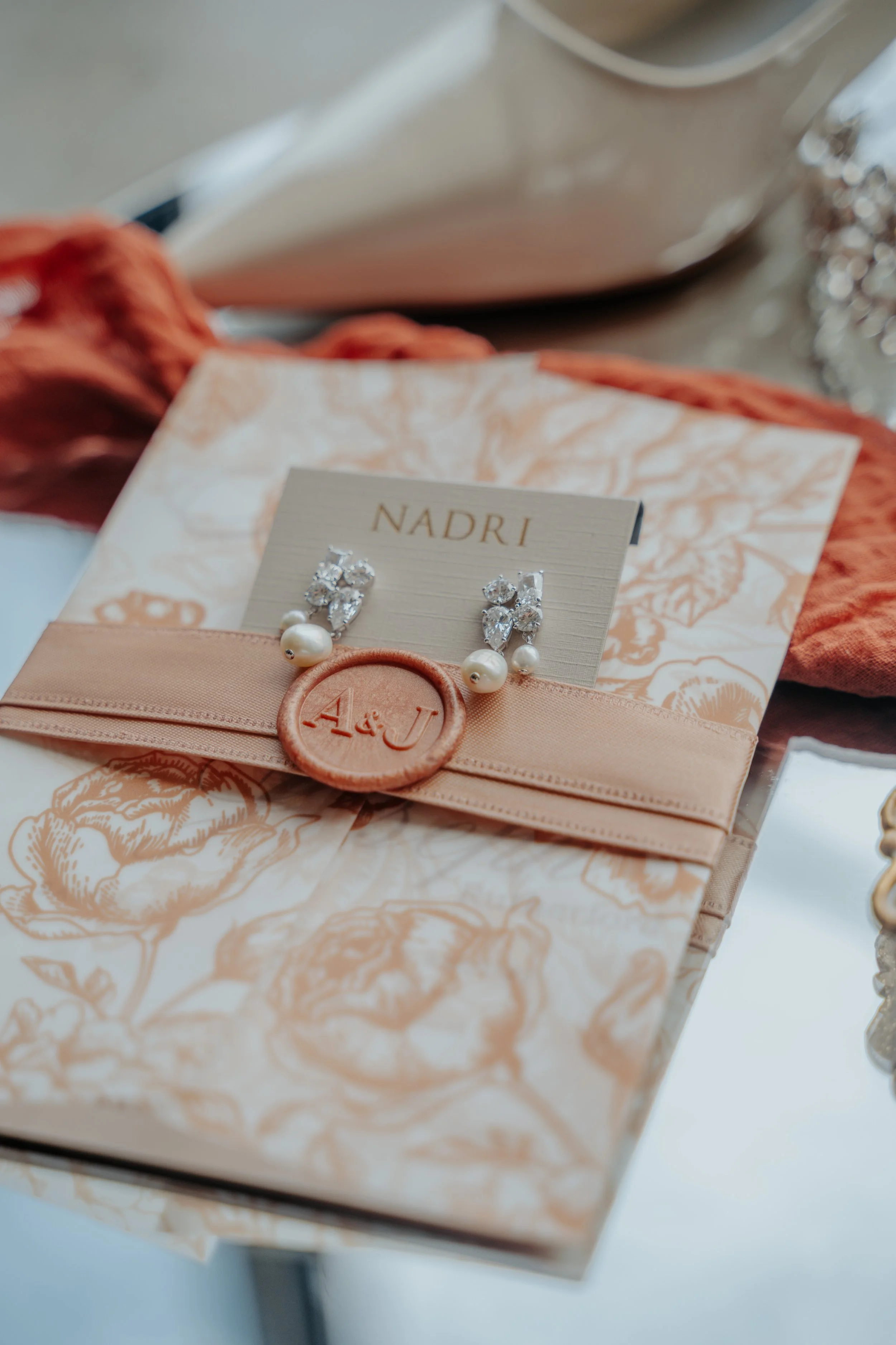Elegant pearl and crystal earrings on a card resting on a floral-patterned paper with a peach-colored ribbon and seal, likely wedding or jewelry accessories.