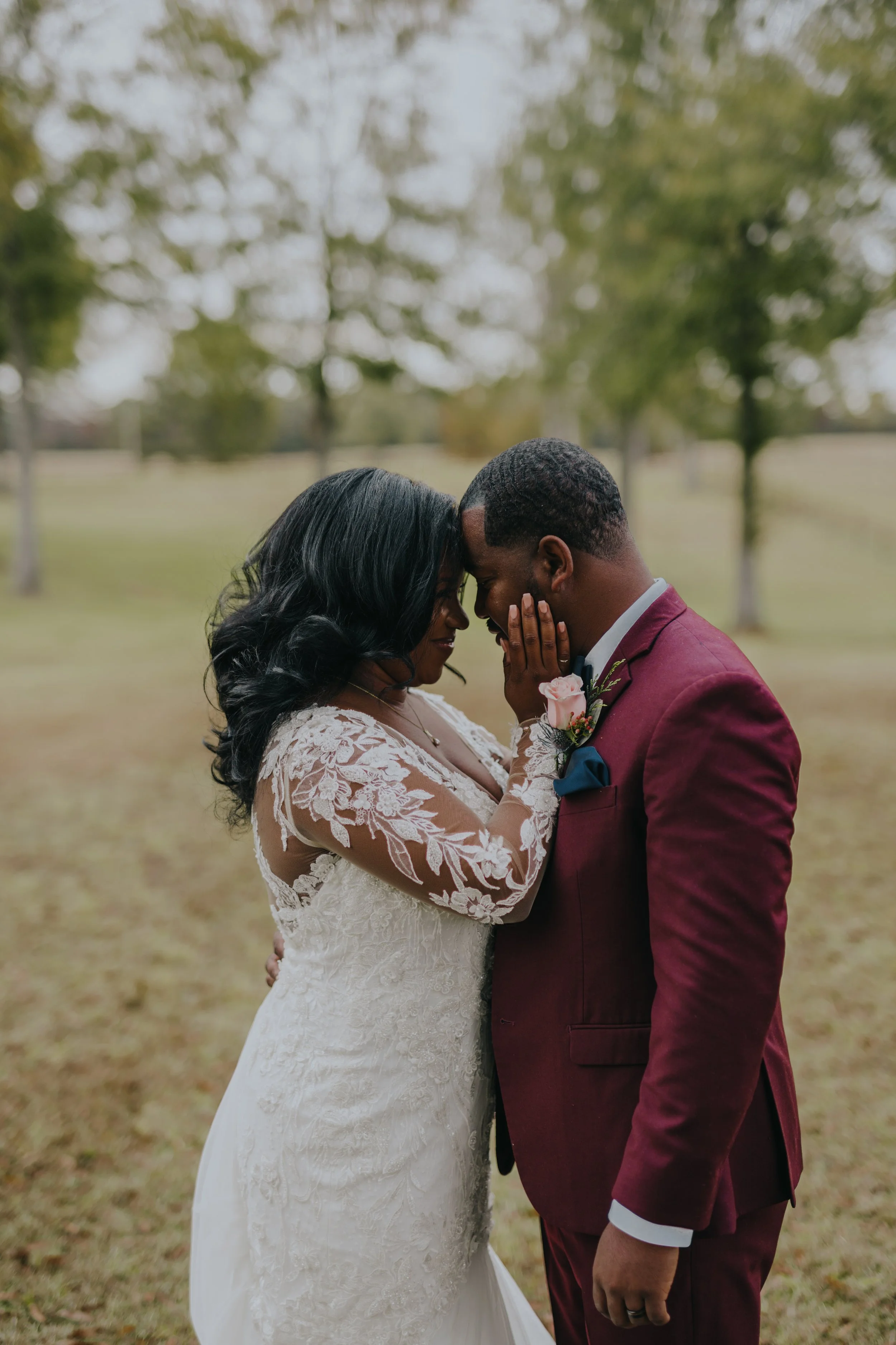 A bride and groom share an intimate moment with their foreheads touching and eyes closed outdoors during daytime, with trees and grass in the background.