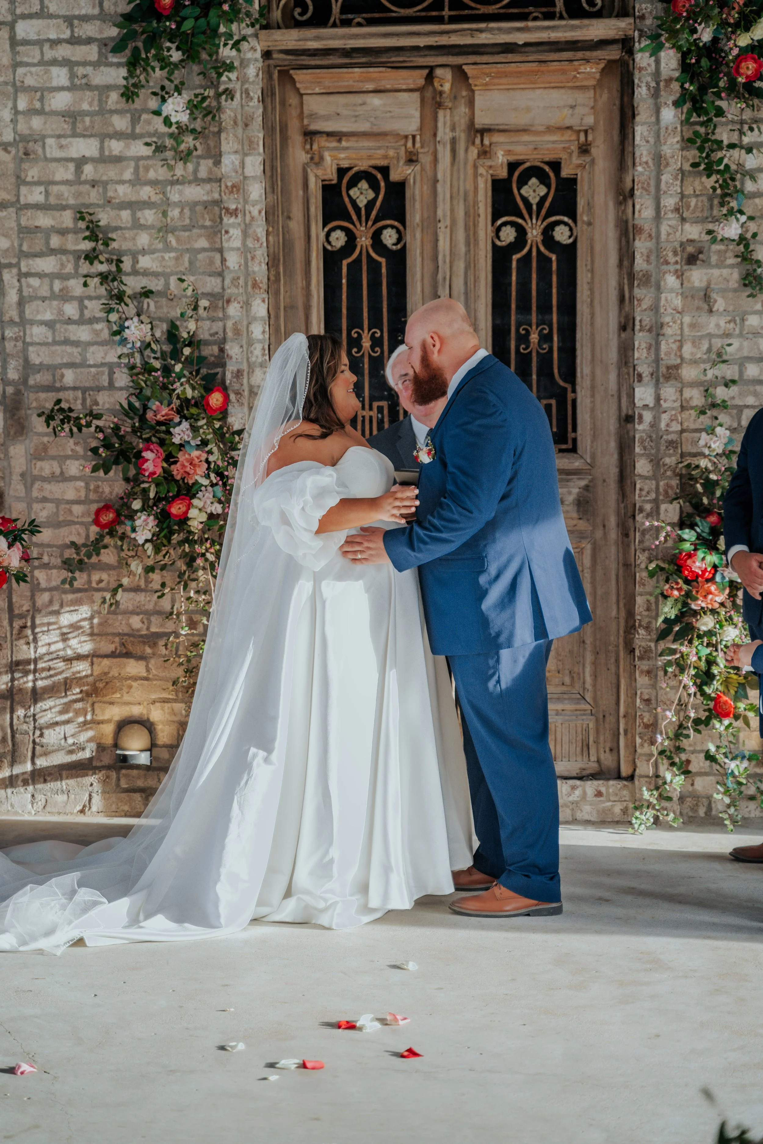 Bride and groom exchanging vows during wedding ceremony with officiant present, floral decorations, and a rustic wooden door in the background.