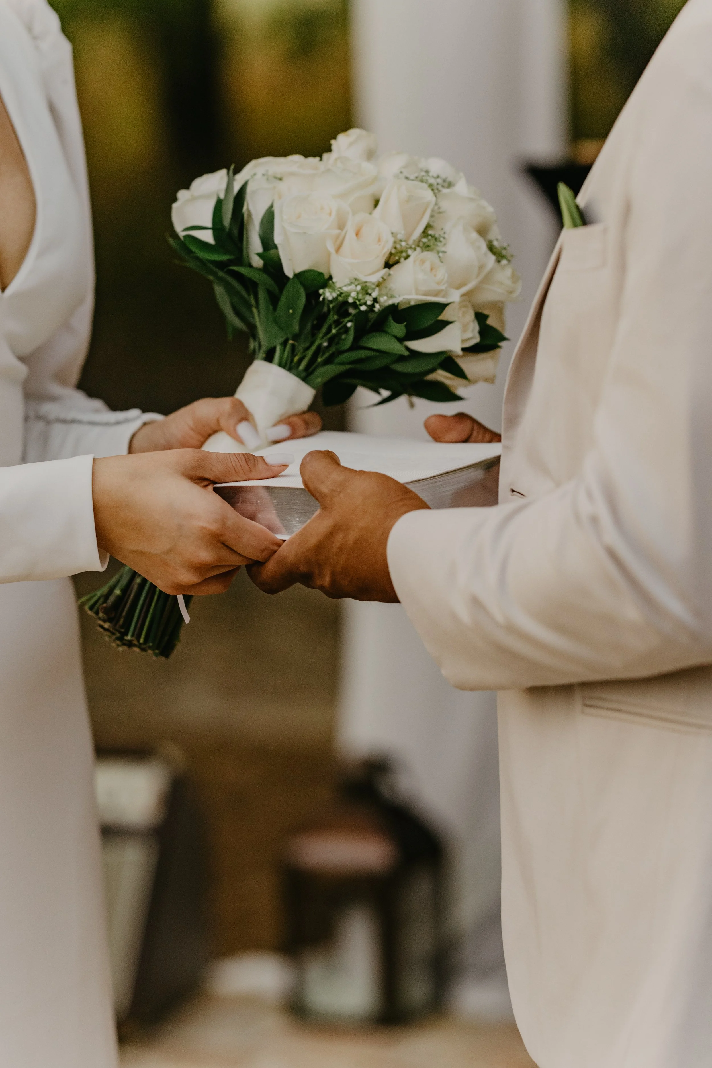 A bride and groom during their wedding ceremony exchanging rings, with the bride holding a bouquet of white roses and greenery.