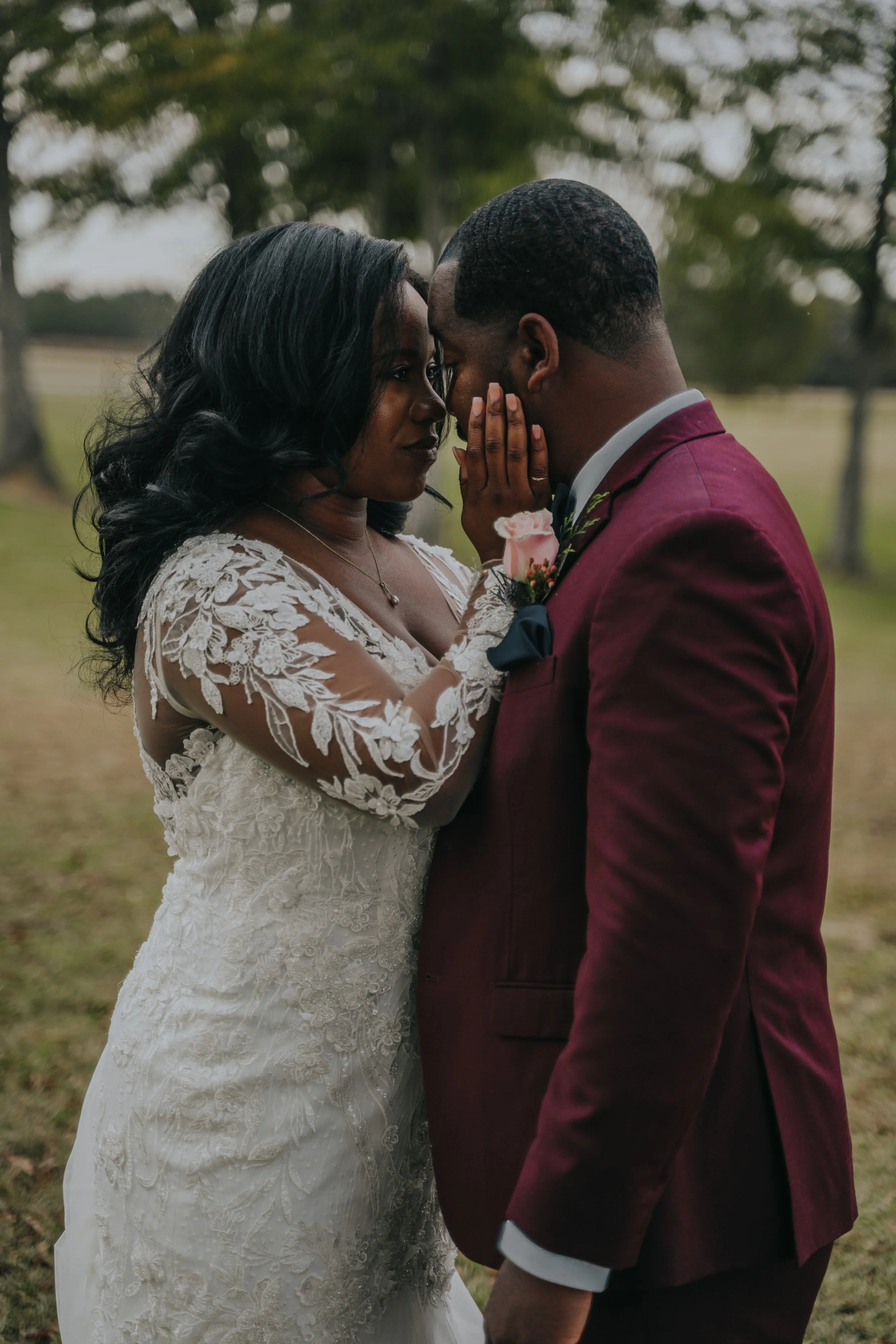 A bride and groom share an intimate moment outdoors, with their foreheads touching and eyes closed, during their wedding. The bride wears a lace wedding dress with floral embroidery, and the groom is dressed in a maroon tuxedo with a boutonniere.
