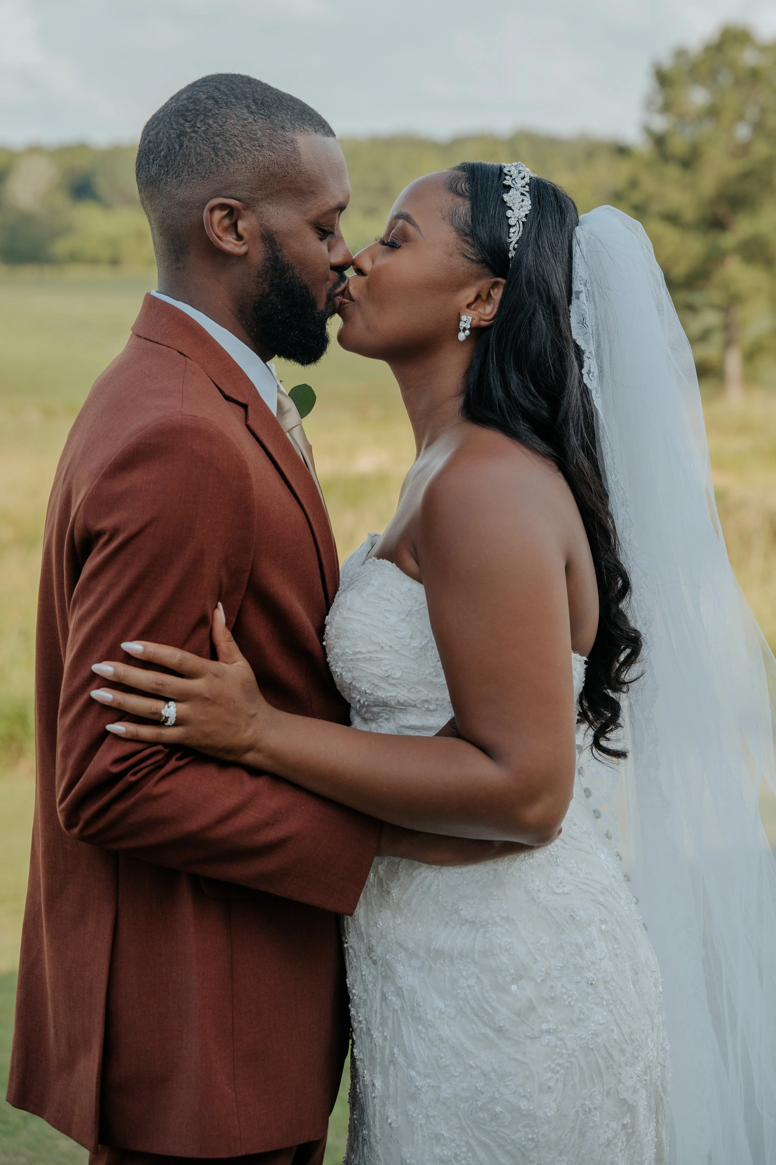 A newlywed couple shares a kiss outdoors, the bride in a white wedding gown with a veil and the groom in a brown suit with a boutonniere, in a green, natural landscape.