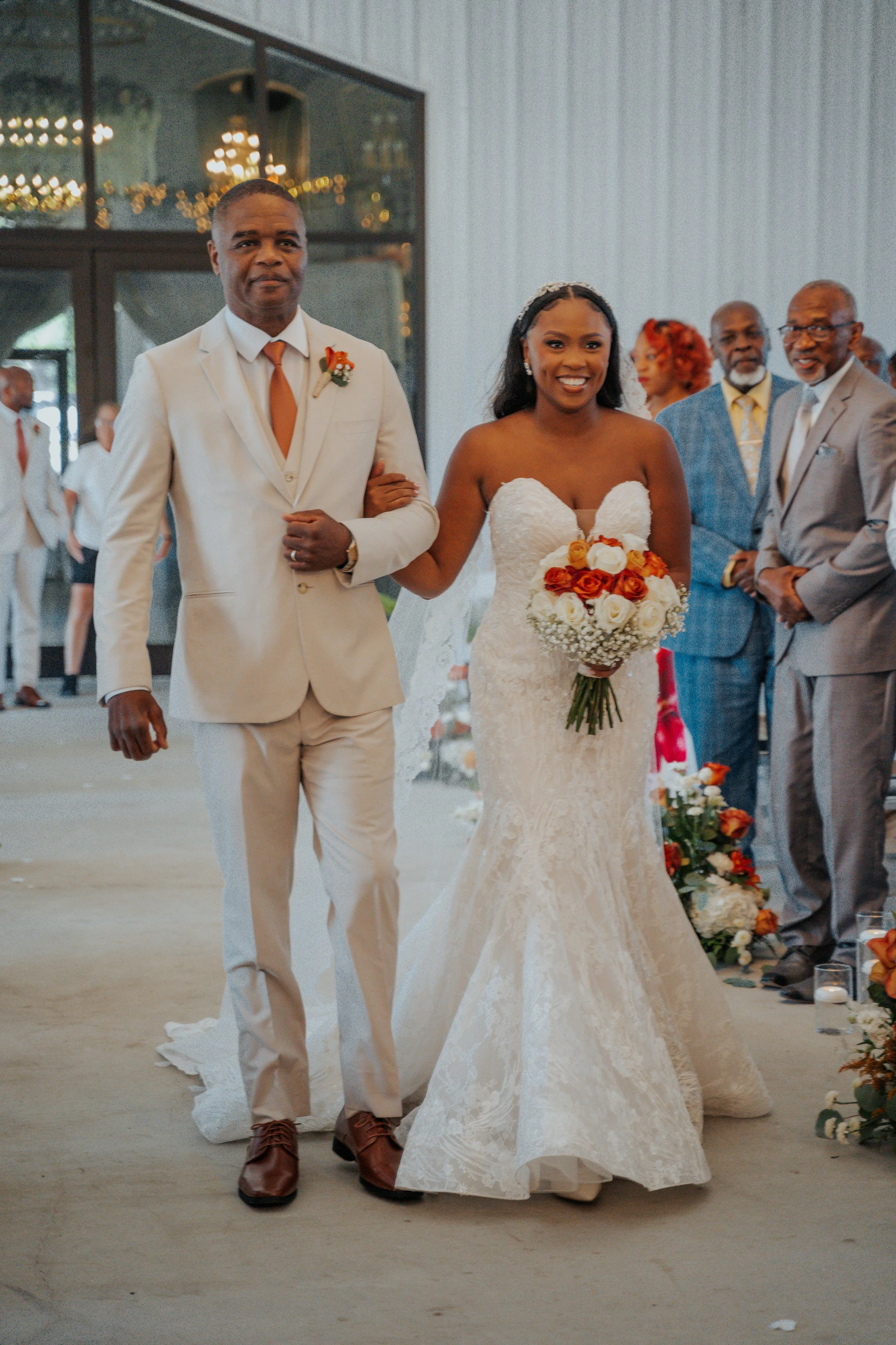 A bride in a white lace wedding dress holding a bouquet of orange and white roses, walking down the aisle arm-in-arm with a man in a white suit, surrounded by smiling guests at a wedding ceremony."