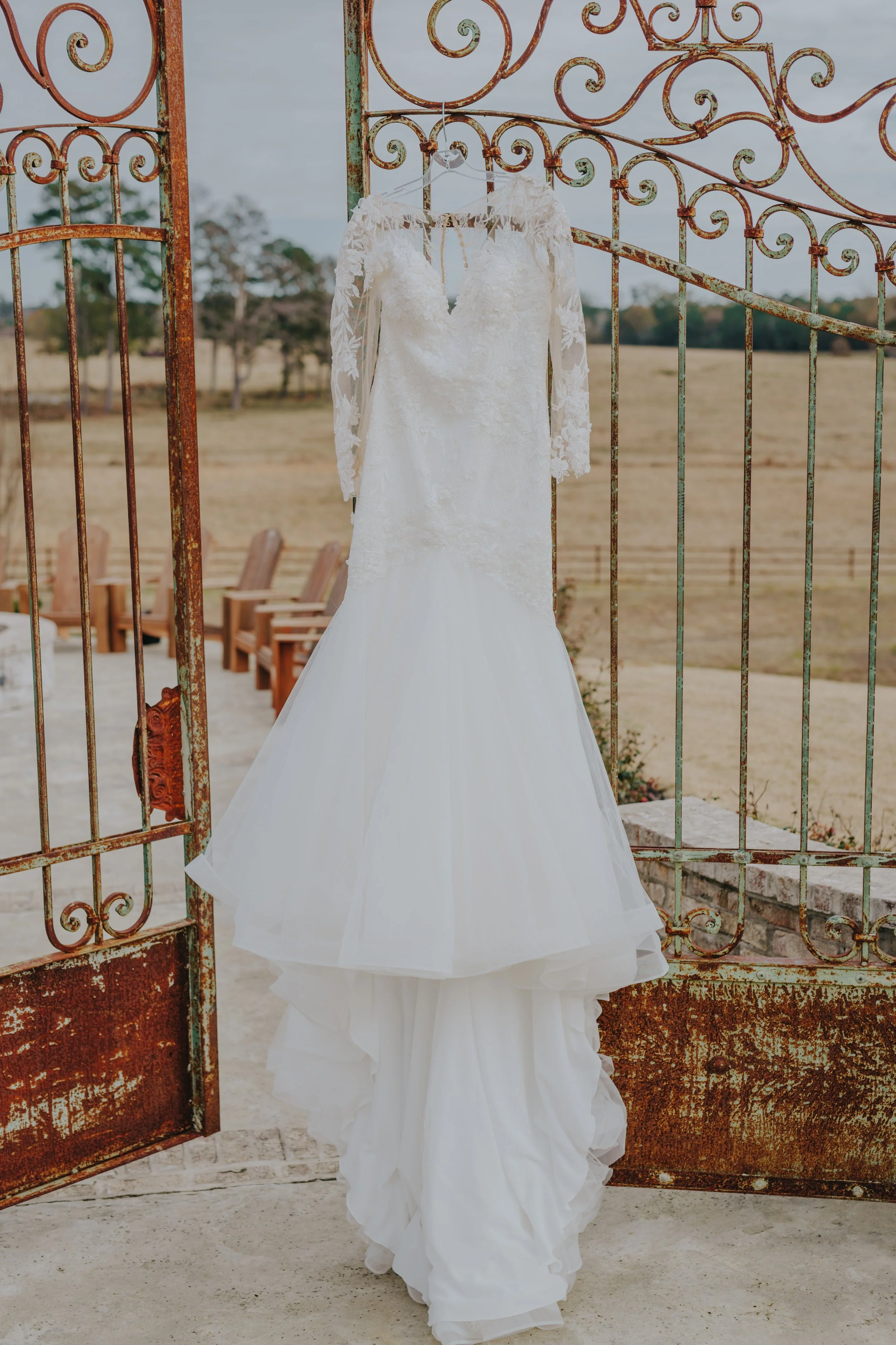 White wedding dress hanging on a hanger outside in front of a rustic open gate overlooking a field.