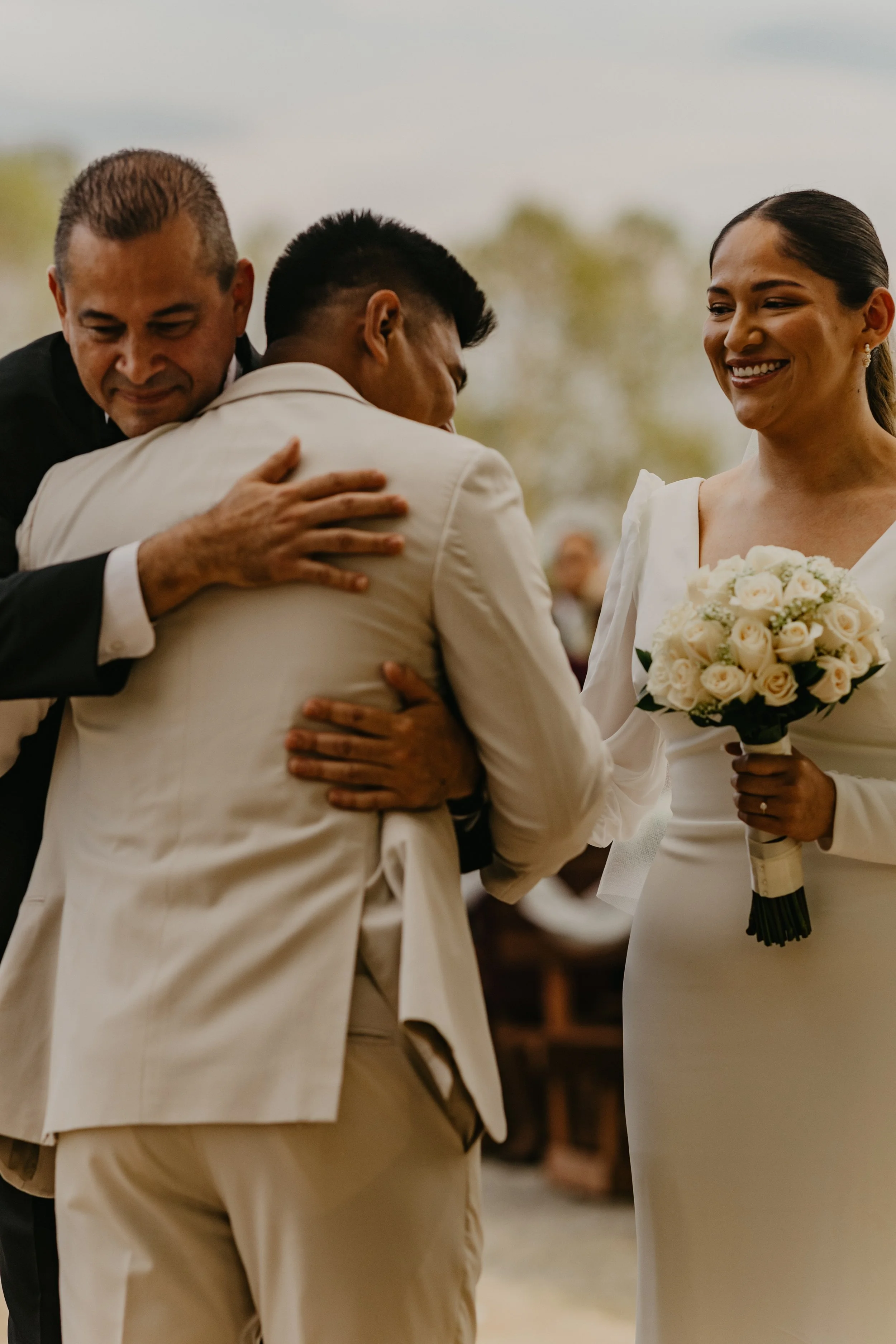 A wedding scene with a woman holding a bouquet of white roses, having a joyful expression, and two men hugging her from either side, one in a black suit and the other in a cream-colored suit, during an outdoor ceremony.