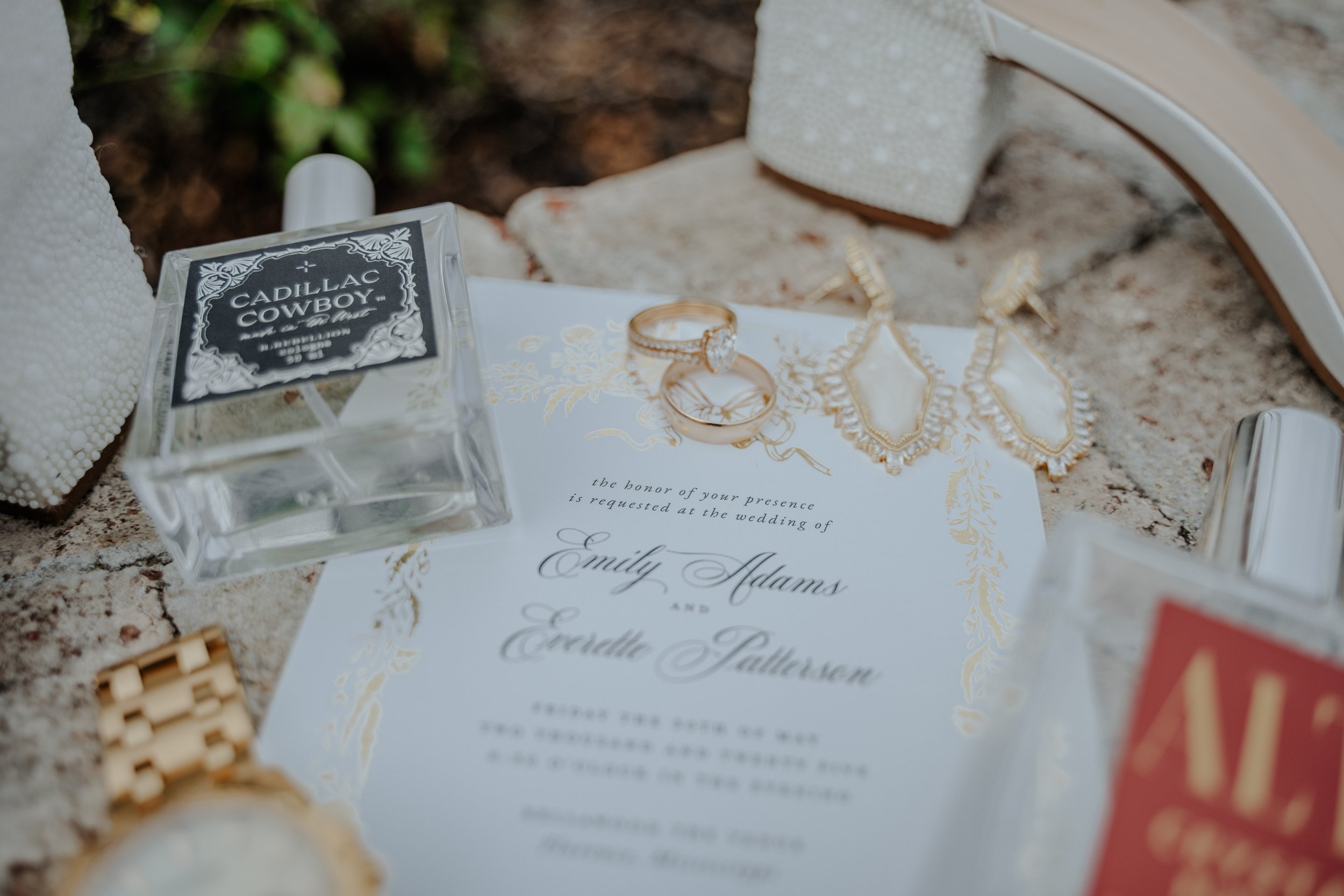 Wedding invitation with gold accents, gold jewelry including rings and earrings, and two bottles of alcohol labeled 'Cadillac Cowboy' and another partially visible bottle.
