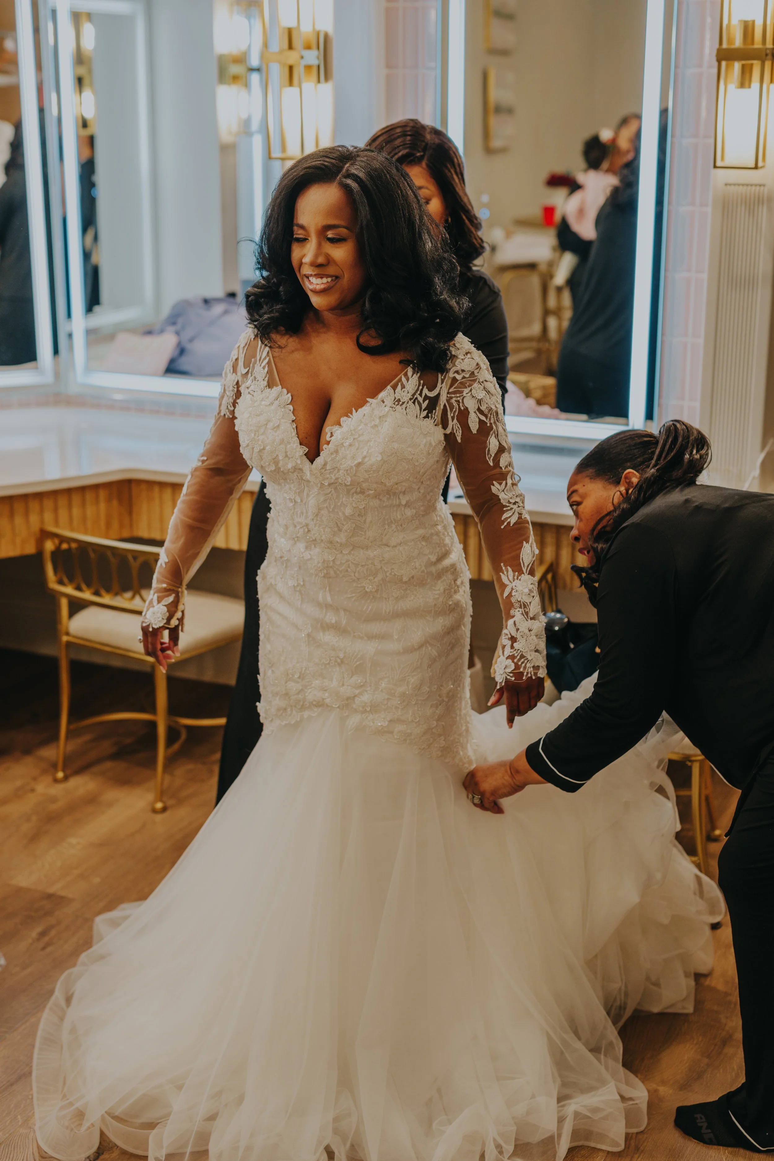 A bride in a lace wedding dress getting ready, assisted by a woman adjusting her gown, with other helpers and a mirror in the background.