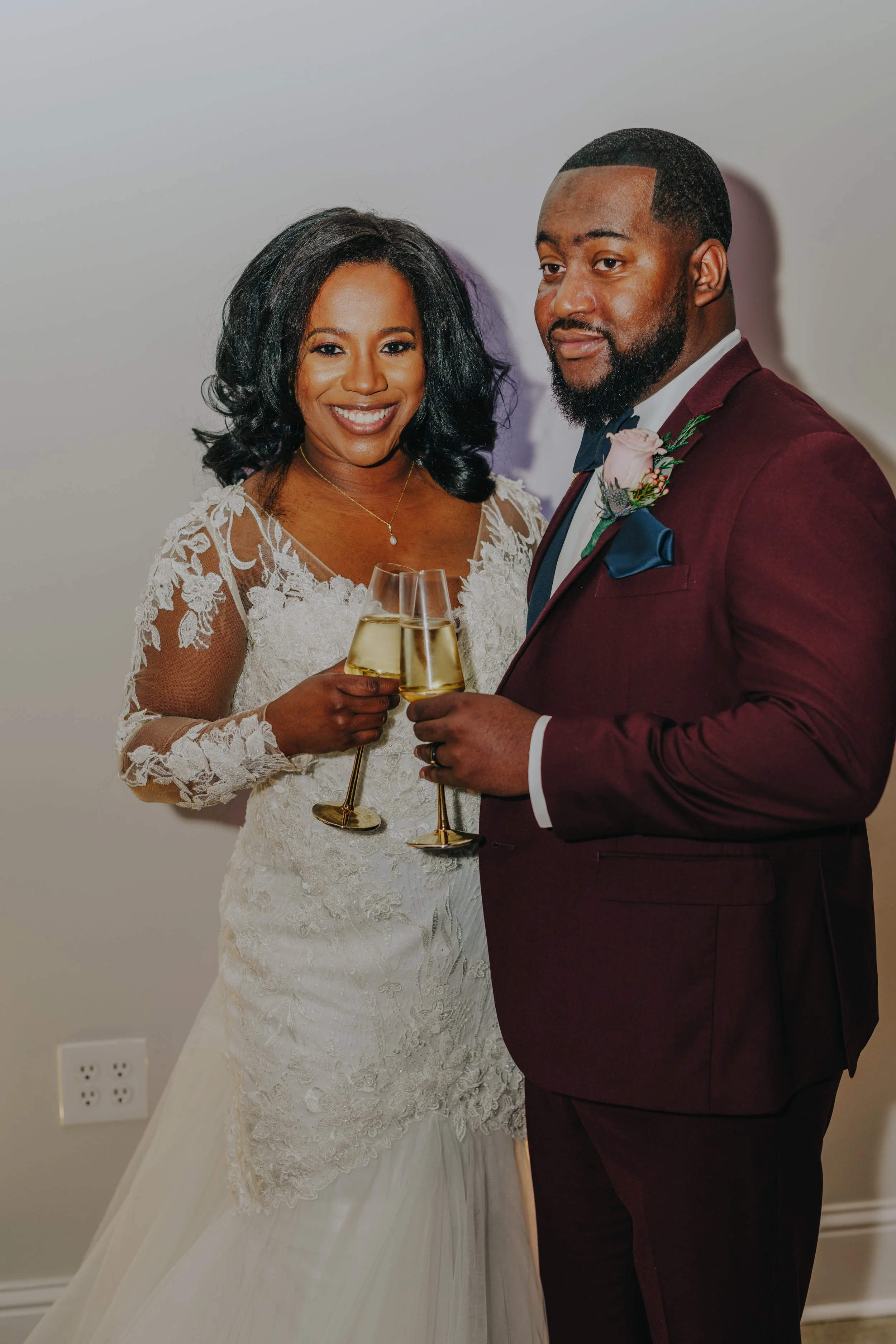 A happy couple at their wedding, holding champagne glasses, dressed in wedding attire, with a plain background.