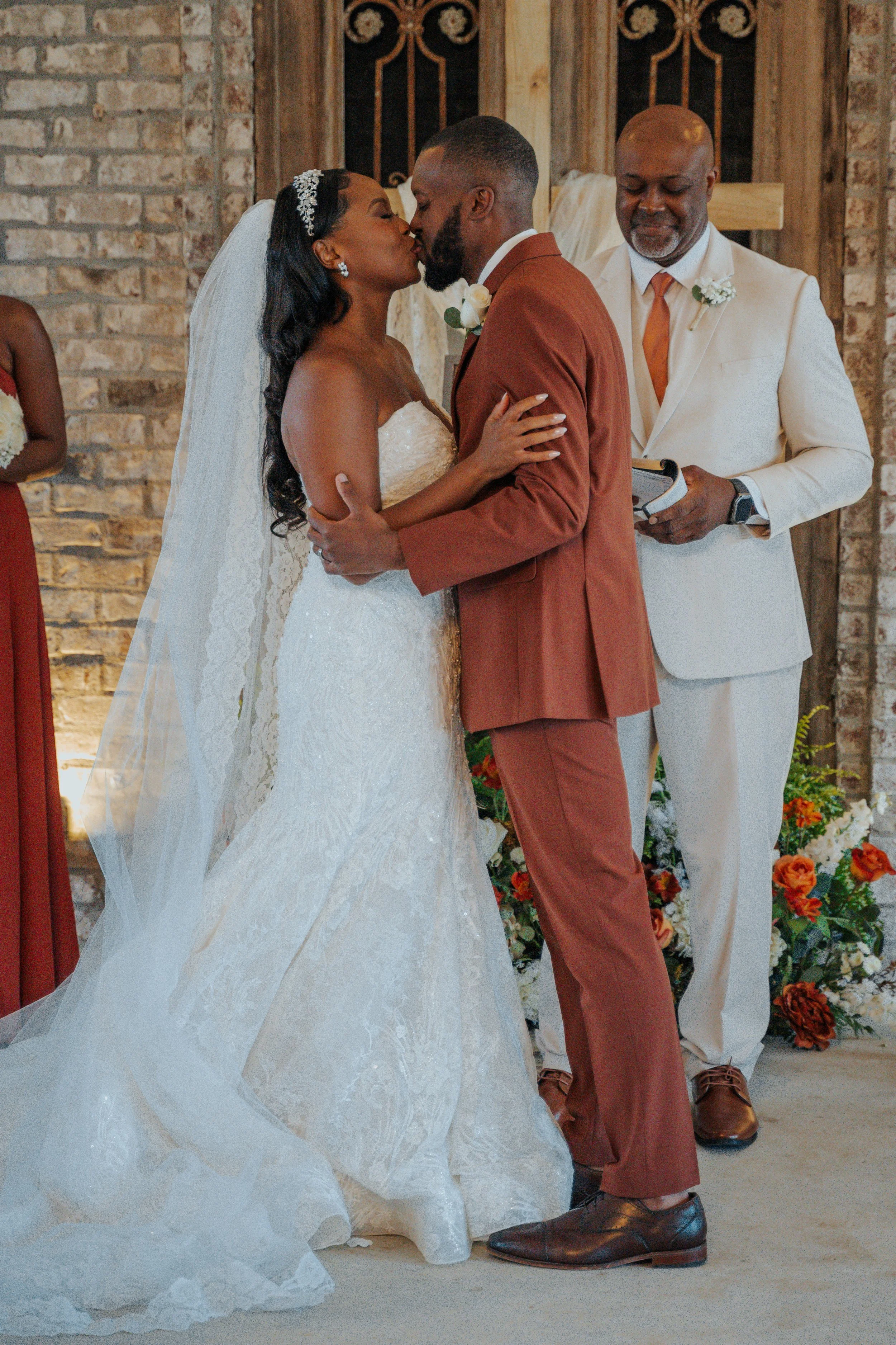 A bride and groom share a kiss during their wedding ceremony, with an officiant standing behind them, in a rustic indoor setting decorated with flowers.