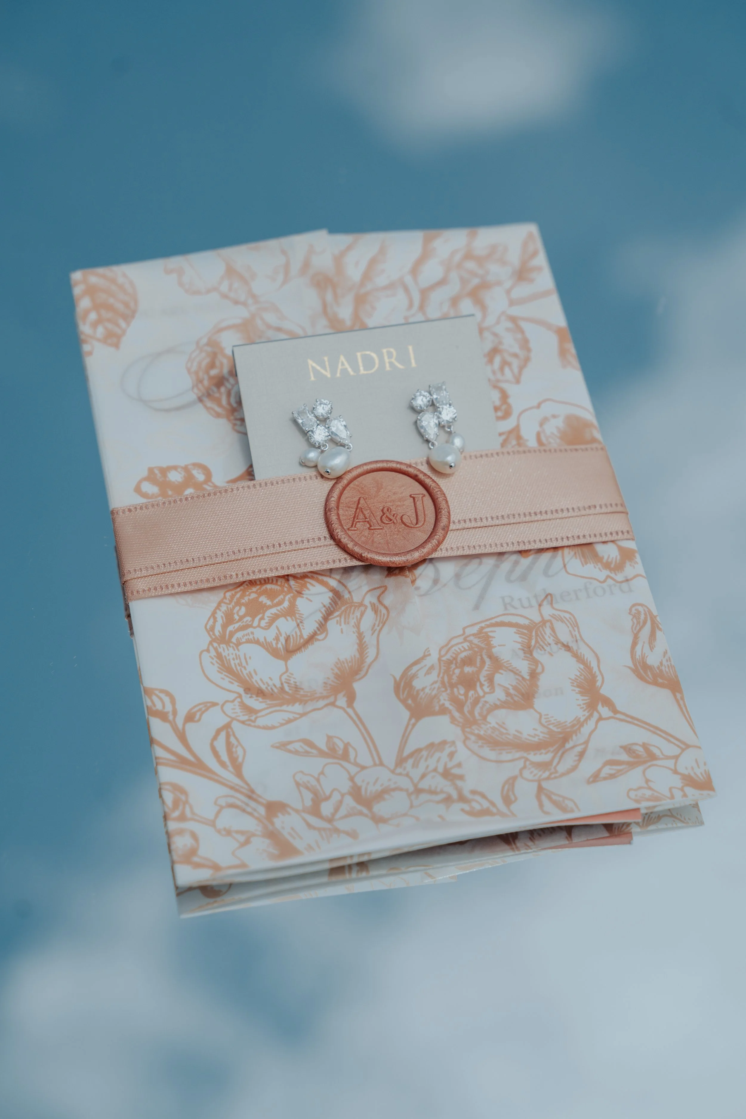 A jewelry box wrapped with a peach ribbon and wax seal, containing pearl earrings, on a floral patterned paper background.