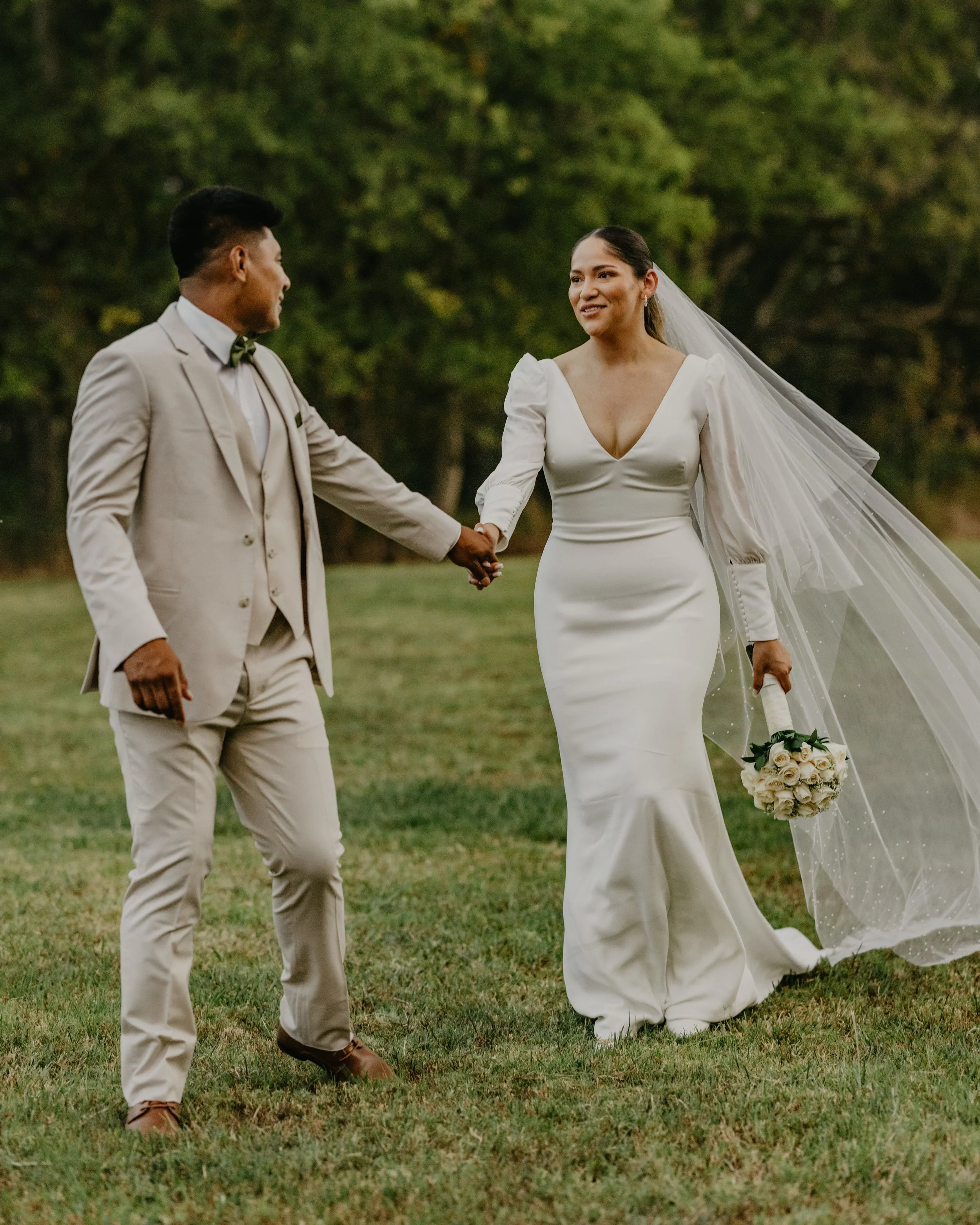 A bride and groom holding hands outdoors on a grassy field with trees in the background, the bride in a white gown with a long veil and holding a bouquet of white roses, the groom in a light beige suit.