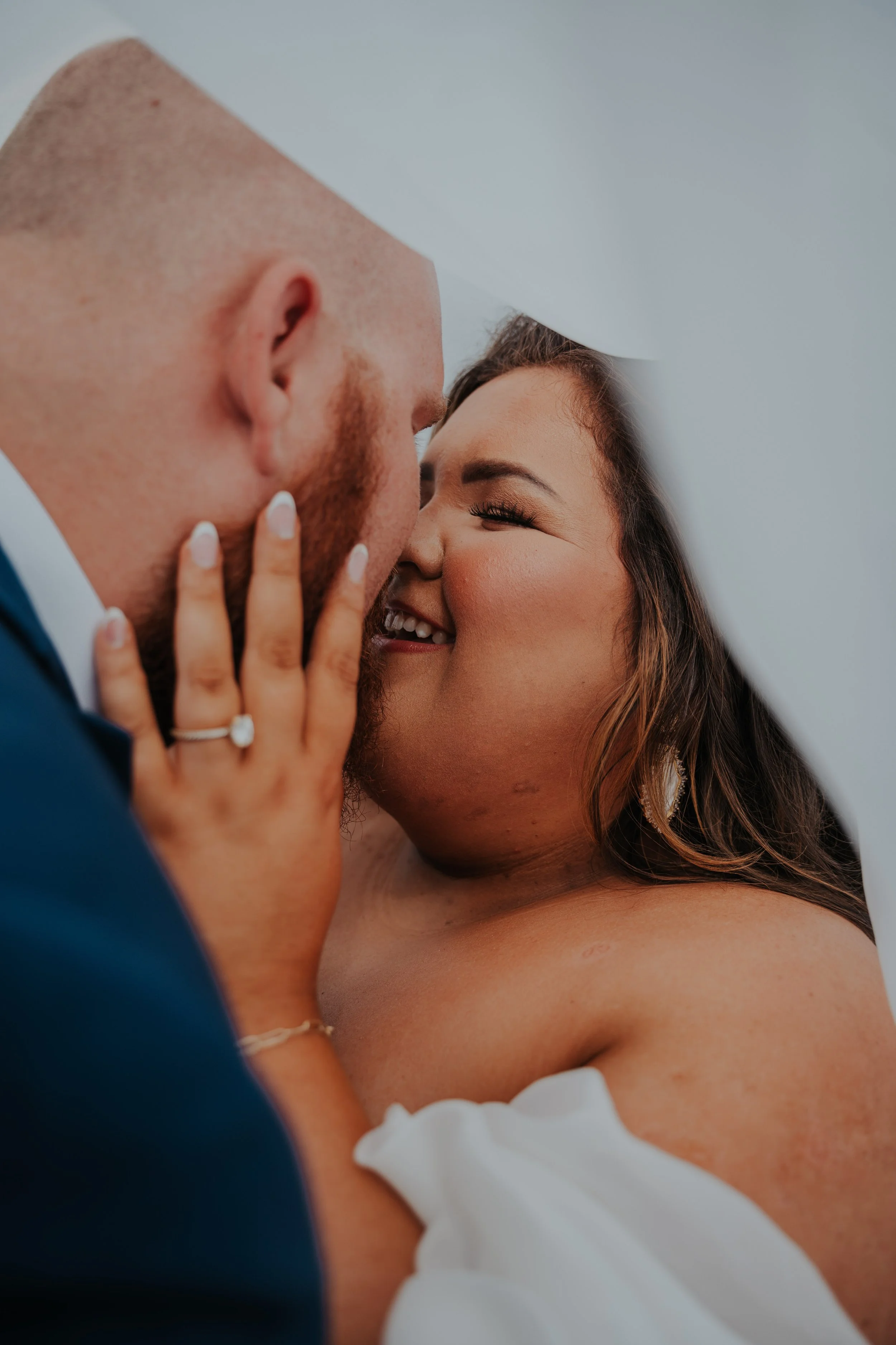 A close-up of a bride and groom sharing an intimate moment under a white veil, with the bride smiling and touching the groom's face.