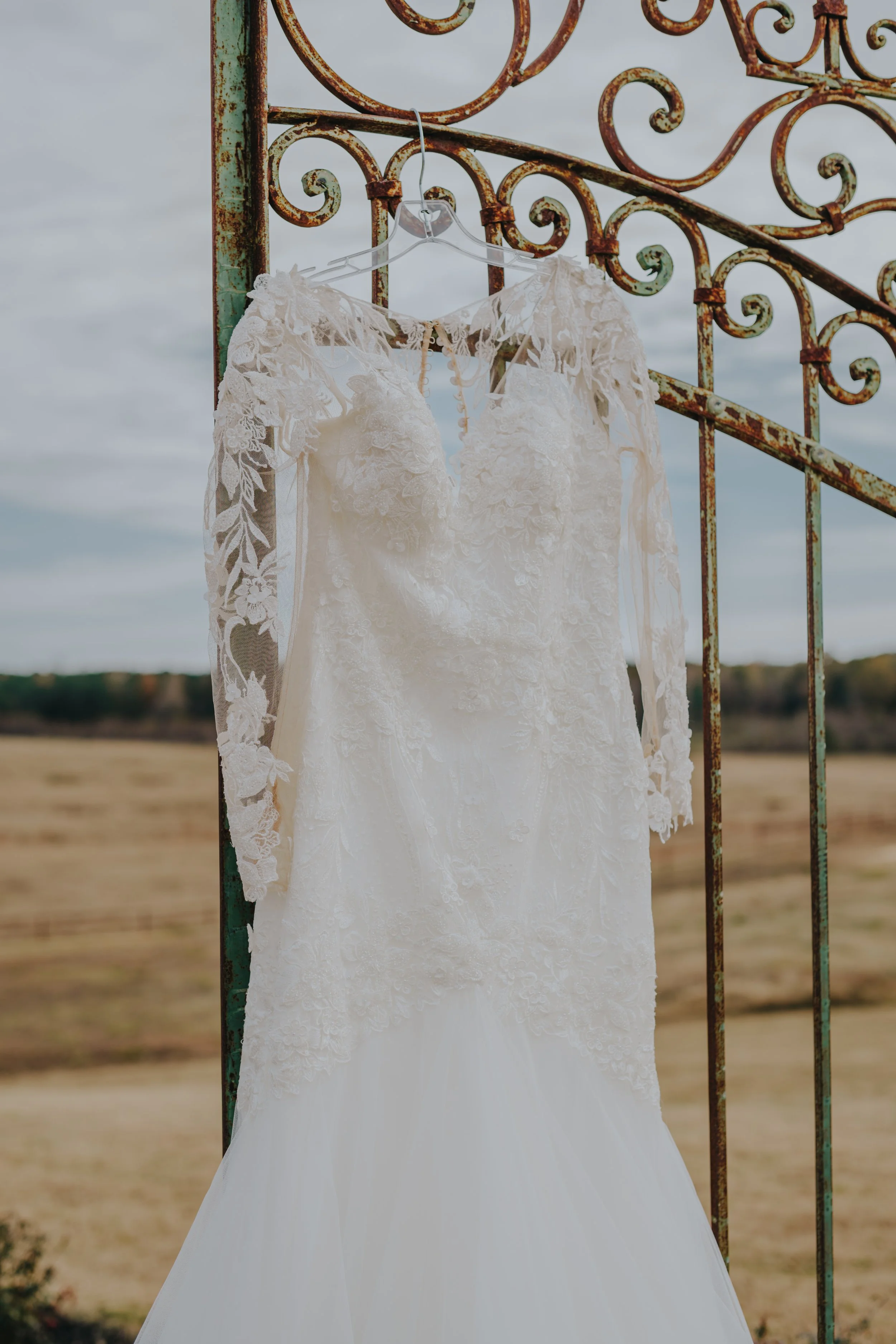 A wedding dress hanging on a clear plastic hanger against a rusty wrought iron gate, with an open field and cloudy sky in the background.
