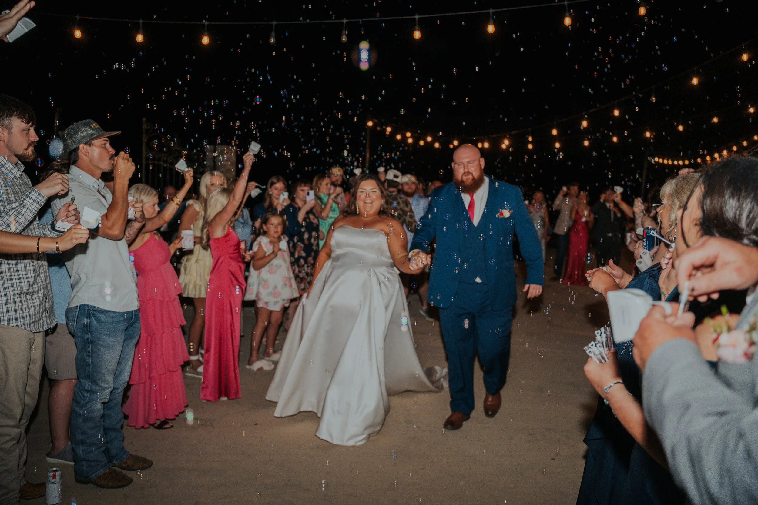 Bride and groom walking through a crowd of guests at night, under string lights, with bubbles floating in the air.