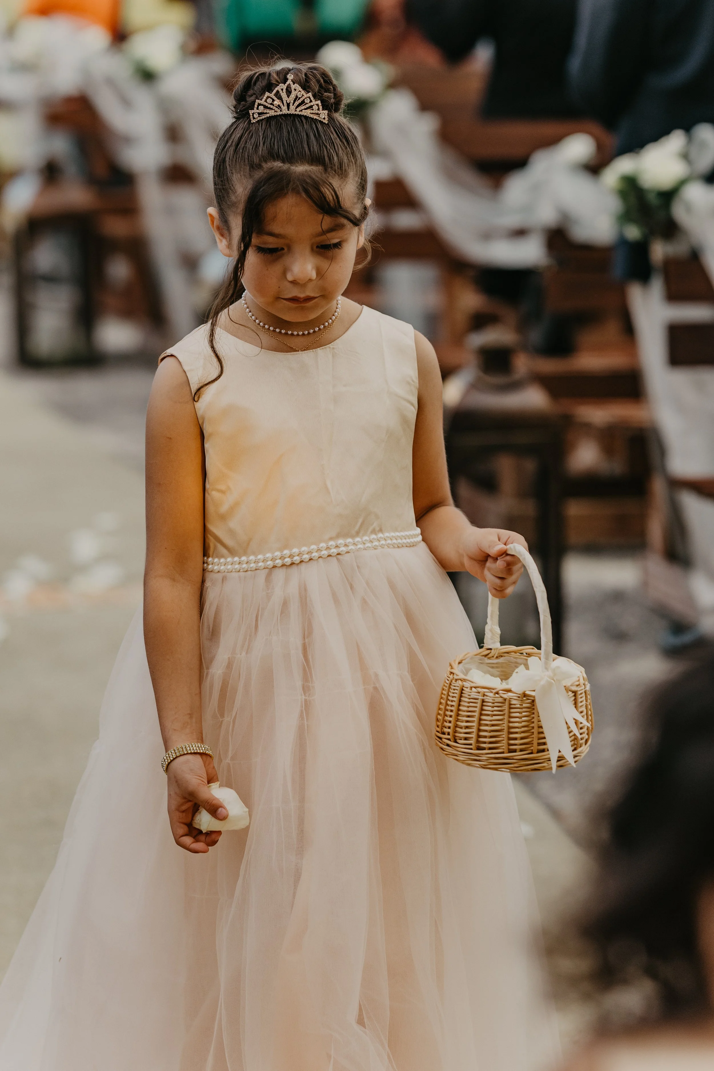 Young girl in a cream-colored dress with pearls, holding a small basket, at a wedding ceremony with pews decorated with white flowers.