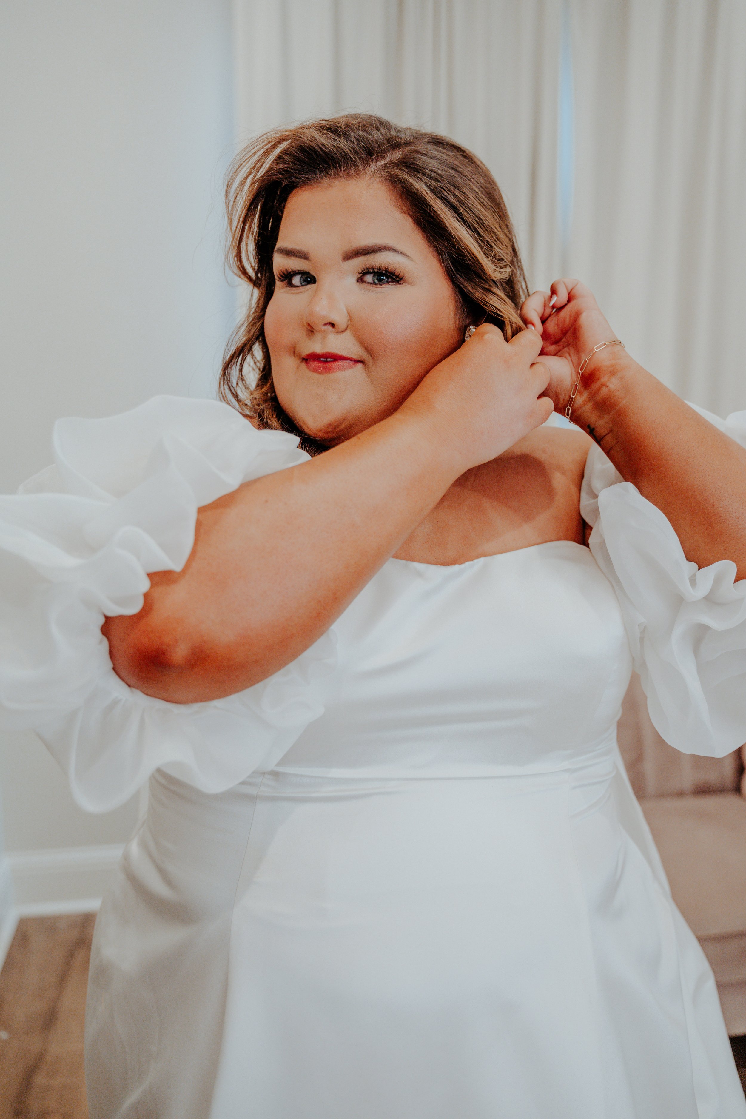 A woman in a white dress adjusting her earring in a room with light-colored curtains.