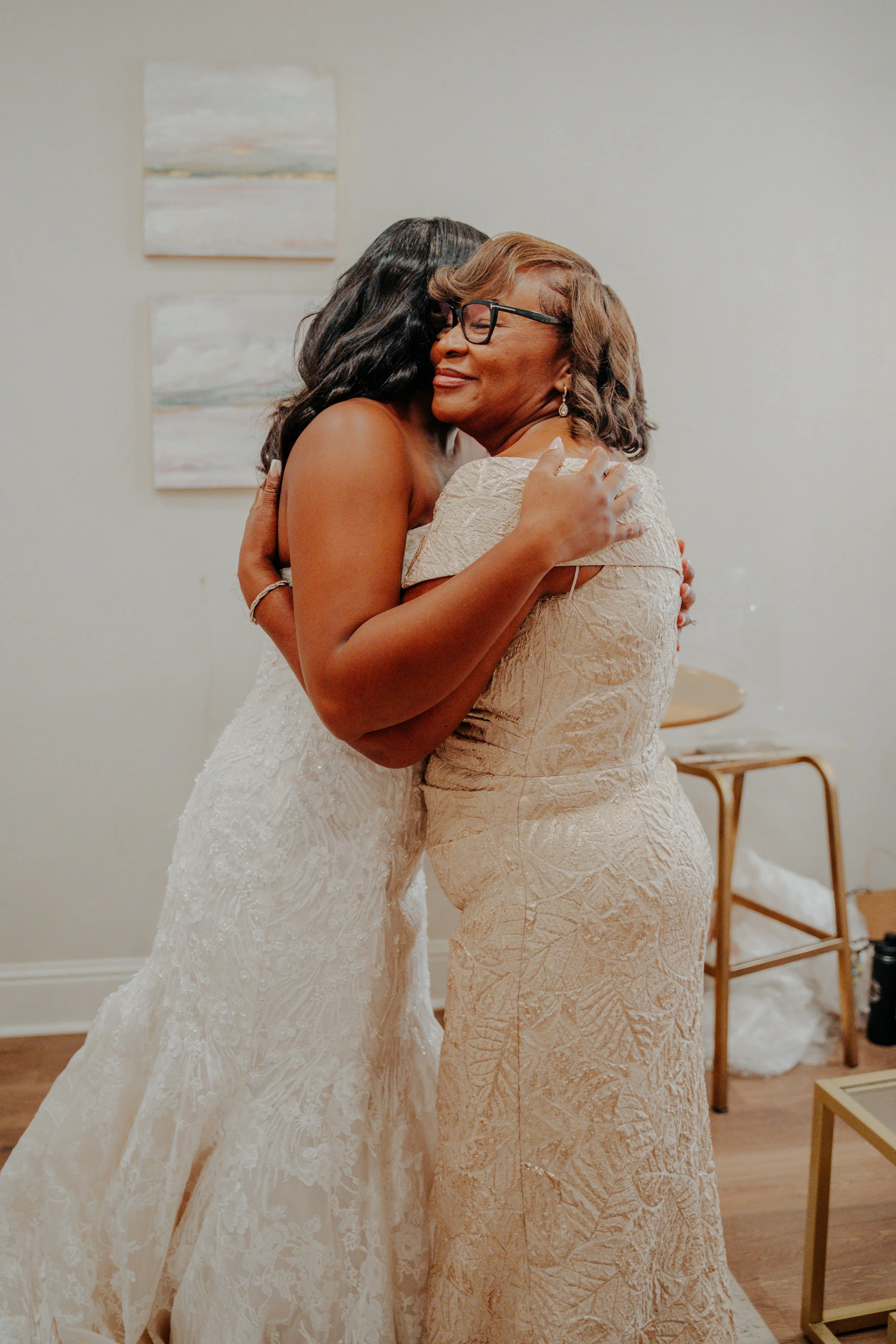 A bride in a wedding gown hugging an older woman in a beige dress.