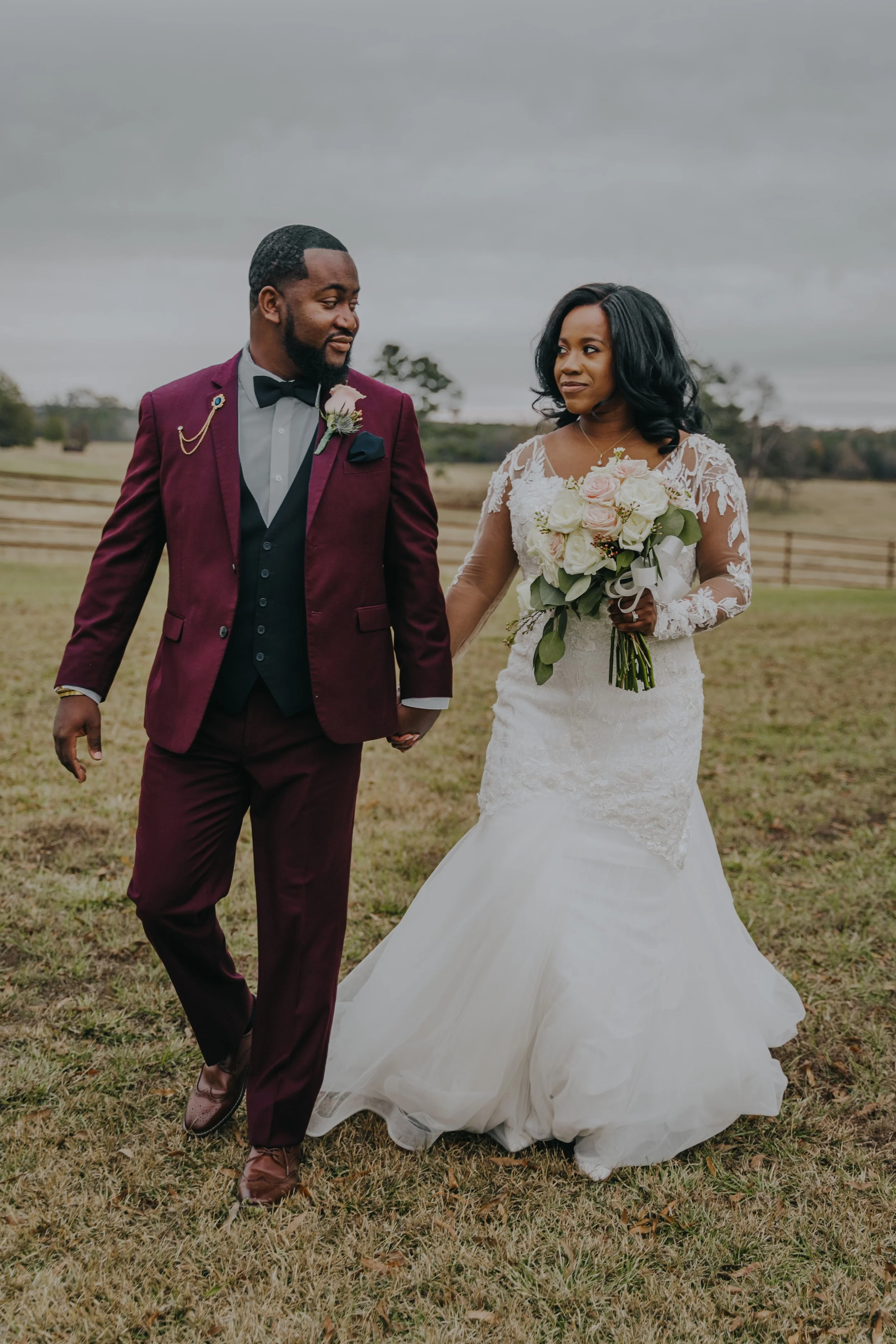 A bride and groom holding hands outdoors on a cloudy day, with the groom in a maroon suit and the bride in a white lace wedding gown holding a bouquet of roses.