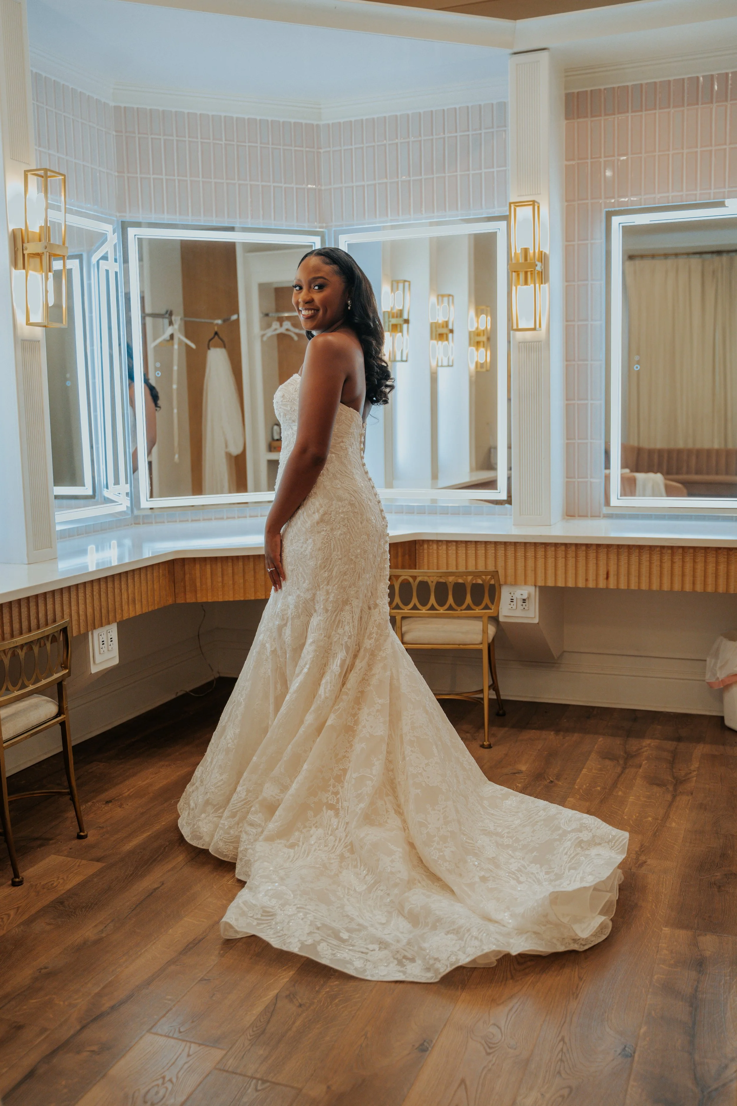 A woman in a strapless wedding dress with a train stands in a dressing room with mirror panels and gold wall sconces, smiling and looking over her shoulder.