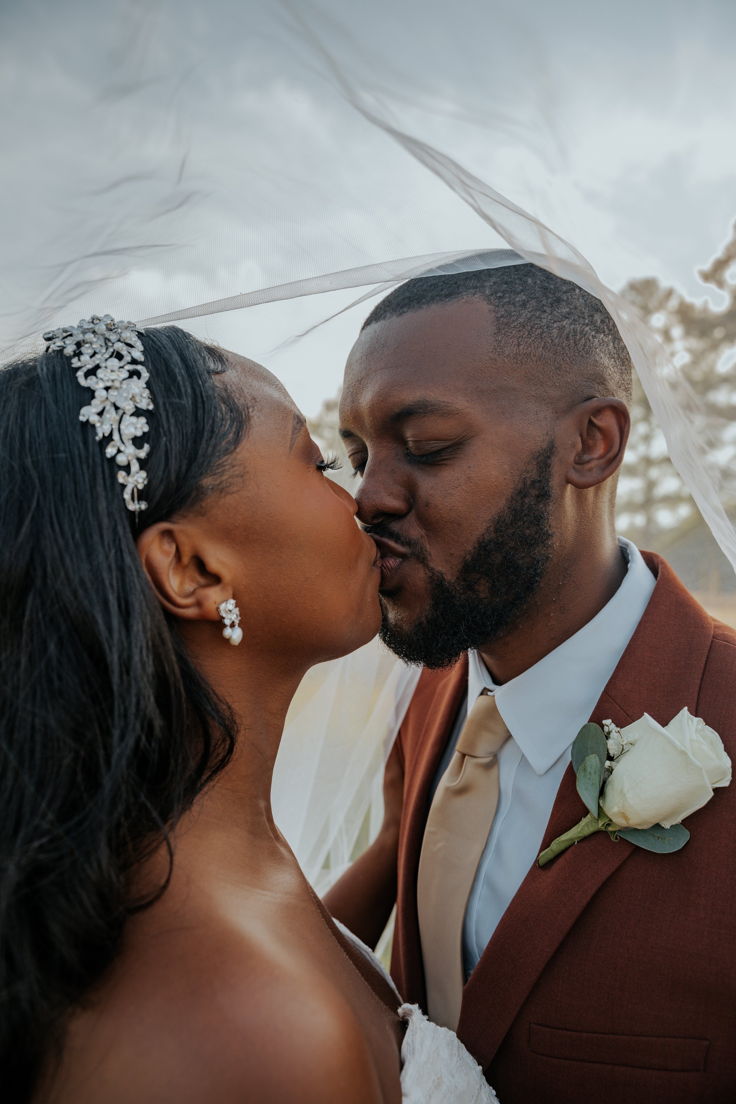 A bride and groom kissing under a veil during their wedding, with the bride wearing pearl earrings and a decorative headband and the groom in a brown suit with a white rose boutonniere.