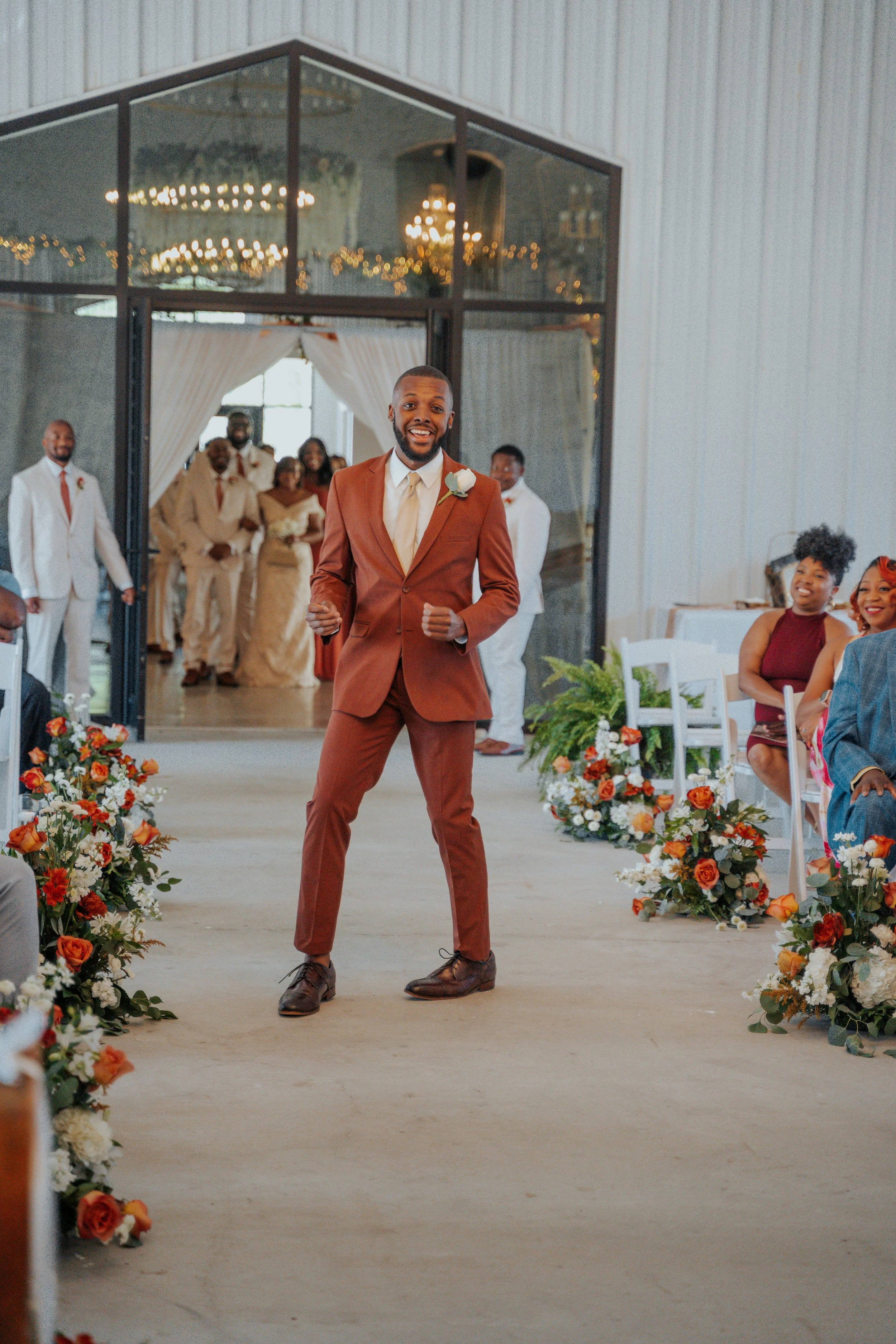 A man in a rust-colored suit stands at a wedding ceremony aisle, smiling with his fists clenched. In the background, wedding guests are seated on either side of the aisle, with floral arrangements on the floor. The ceremony is taking place inside a r