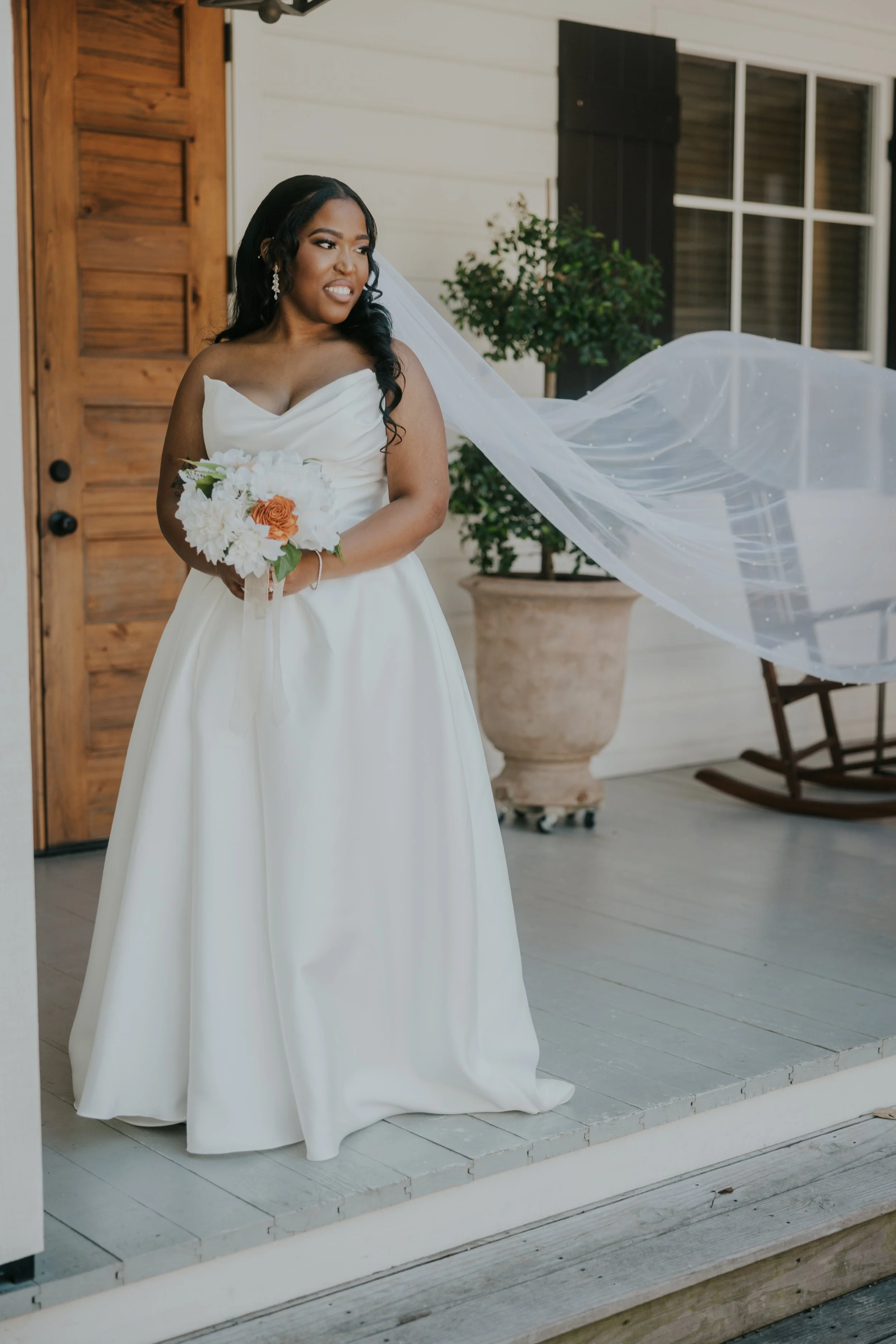 Bride in a white wedding gown holding a bouquet of white and orange flowers, standing on a porch with a wooden door and a rocking chair in the background.