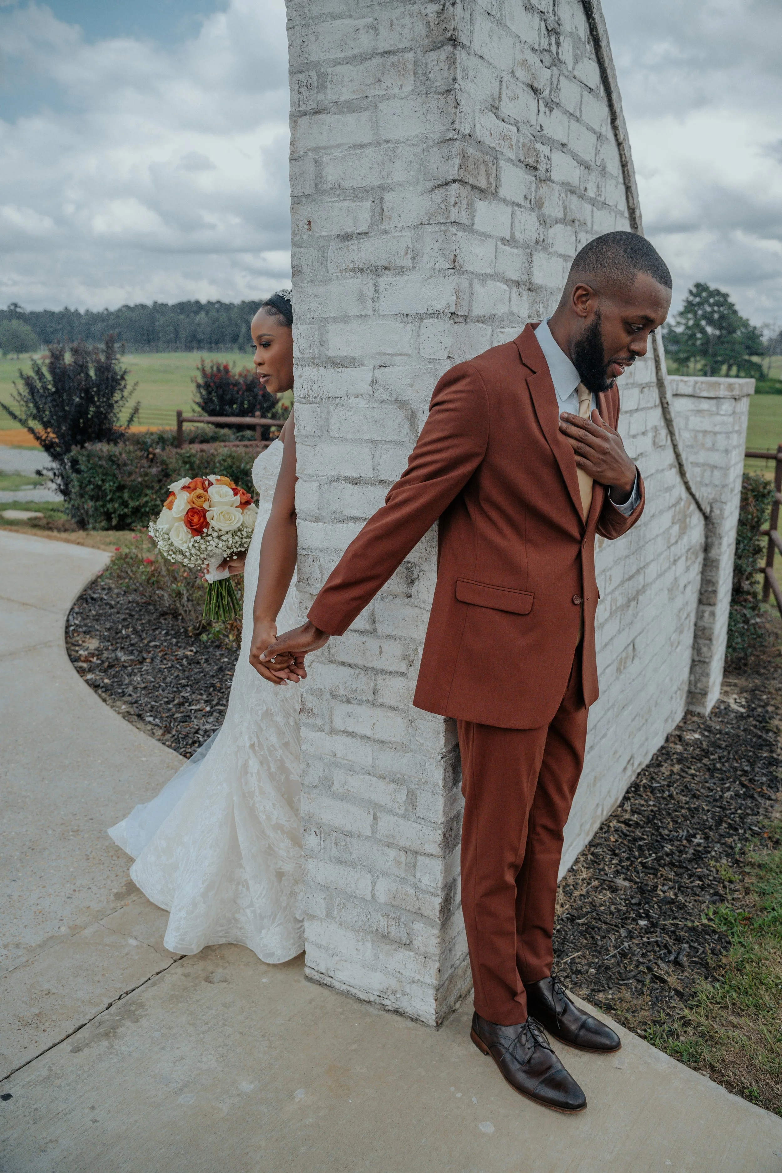 A bride and groom are standing back to back outside, holding hands behind a brick wall. The bride is in a white wedding dress holding a bouquet, and the groom is in a brown suit touching his chest with his other hand.