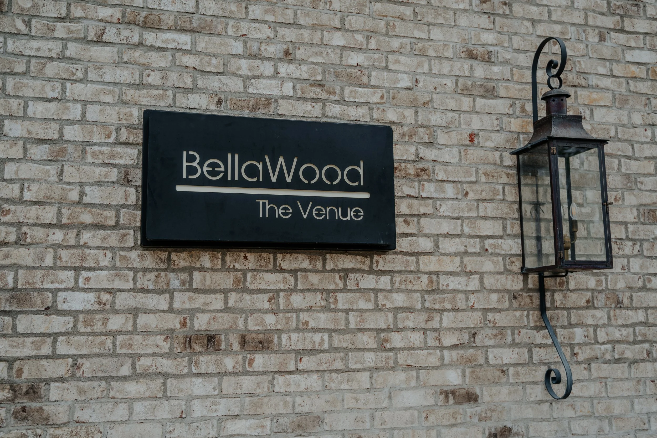 A black sign reading 'BellaWood The Venue' mounted on a brick wall, with a vintage-style lantern to its right.