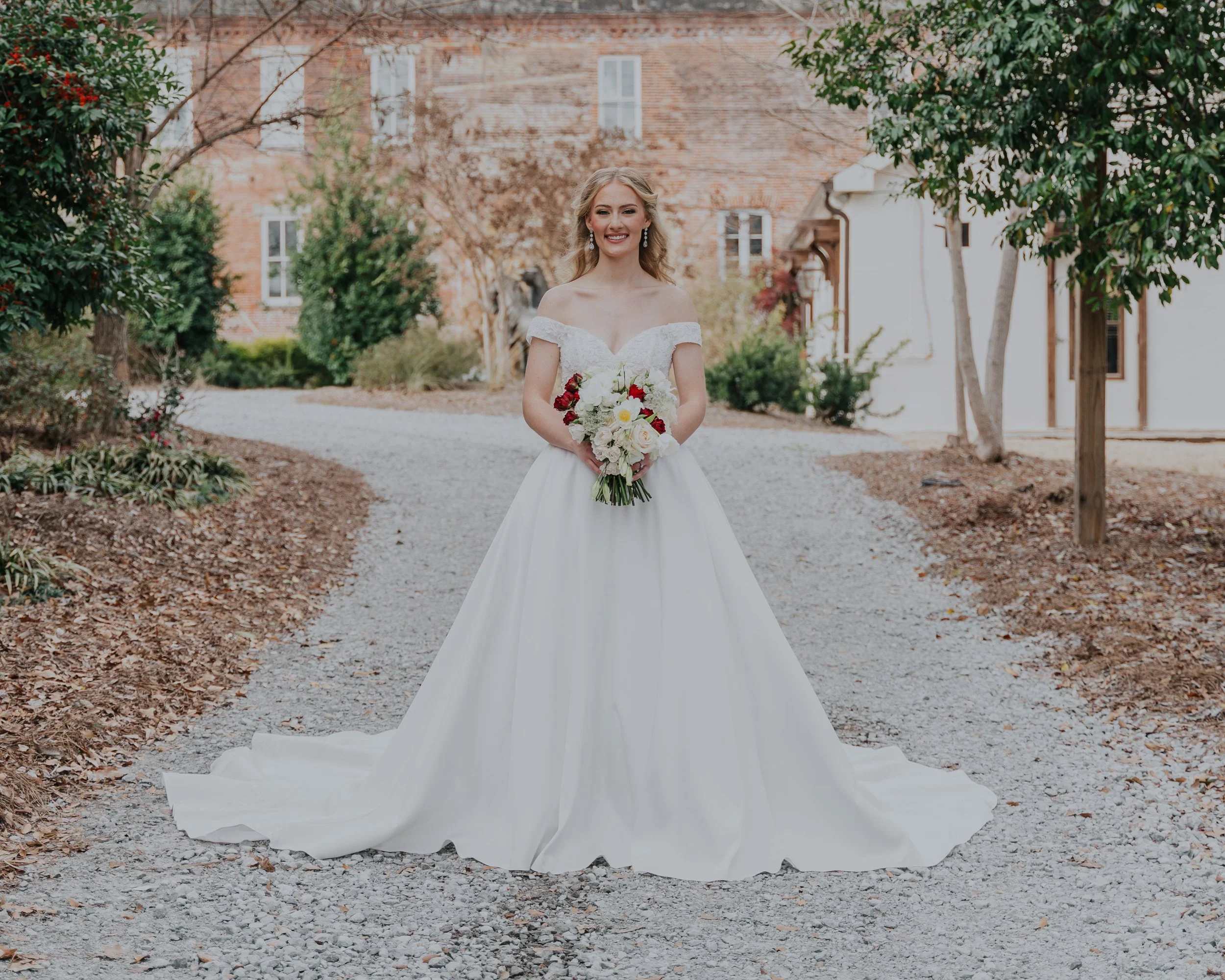 A smiling bride standing outdoors on a gravel path, holding a bouquet of white and red flowers, wearing a white off-shoulder wedding gown with a train, surrounded by trees and buildings.