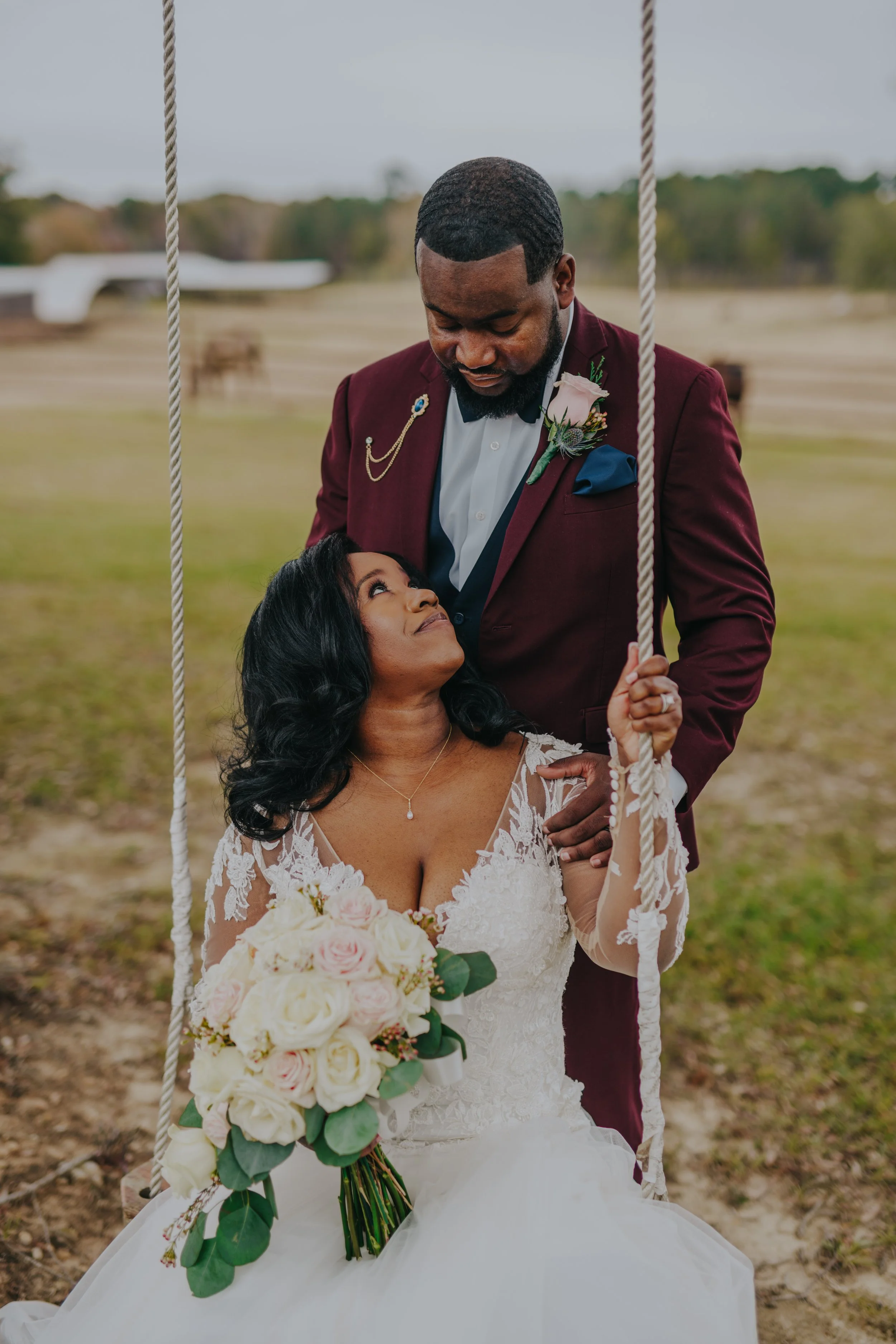 A bride sitting on a swing holding a bouquet, looking up at a groom standing next to her, outdoors in a field.
