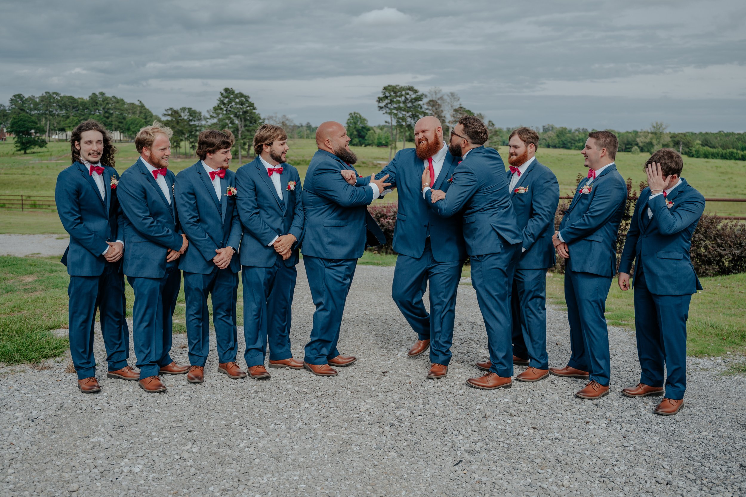 A group of men in blue suits and red bow ties standing outdoors on a gravel path, engaging in playful interactions with some laughing and covering their faces, with a scenic green landscape and cloudy sky in the background.