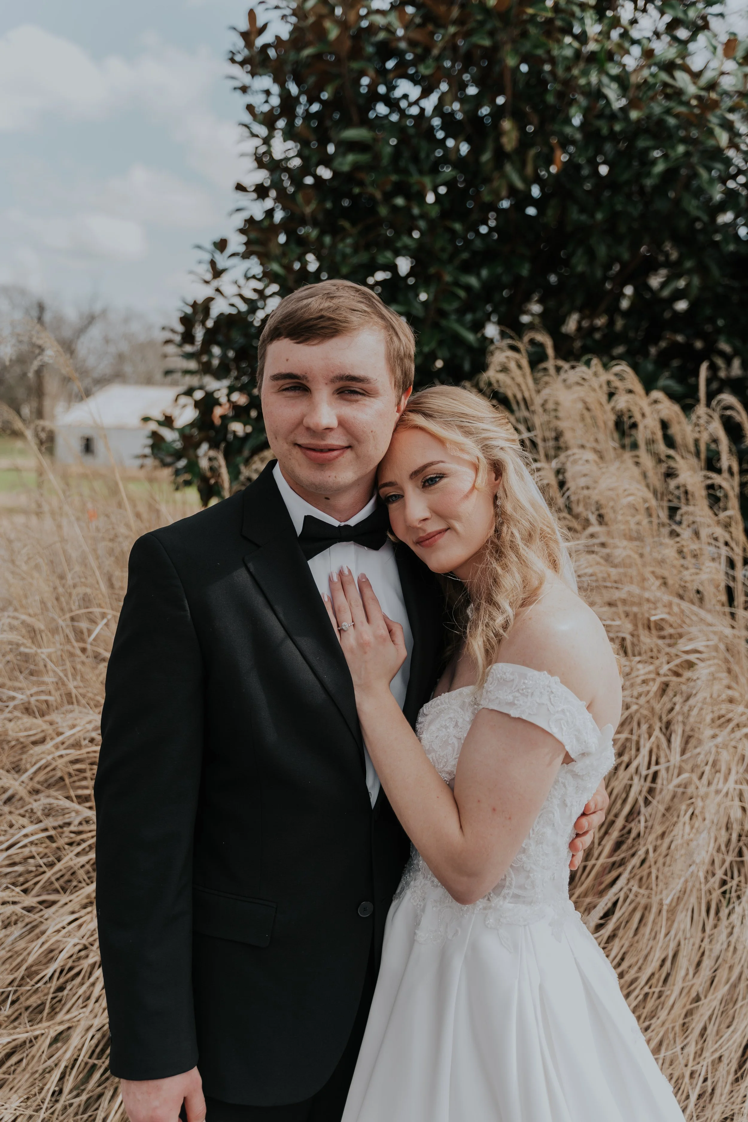 A bride and groom embracing outdoors, with dry grass and a large tree in the background.
