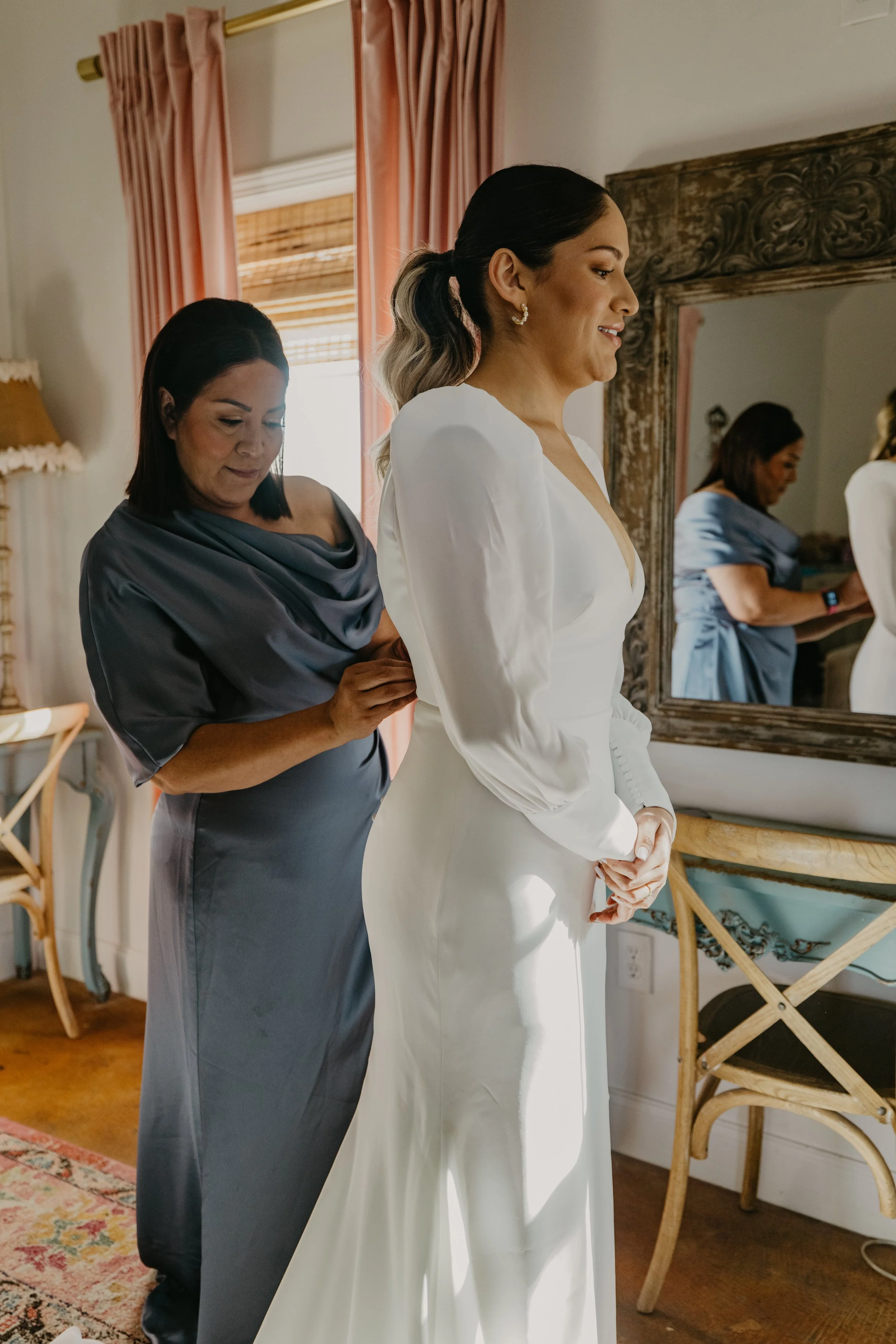 A bride in a white wedding gown is being helped with her dress by a woman in a silver satin dress in a room with pink curtains and a wooden mirror.