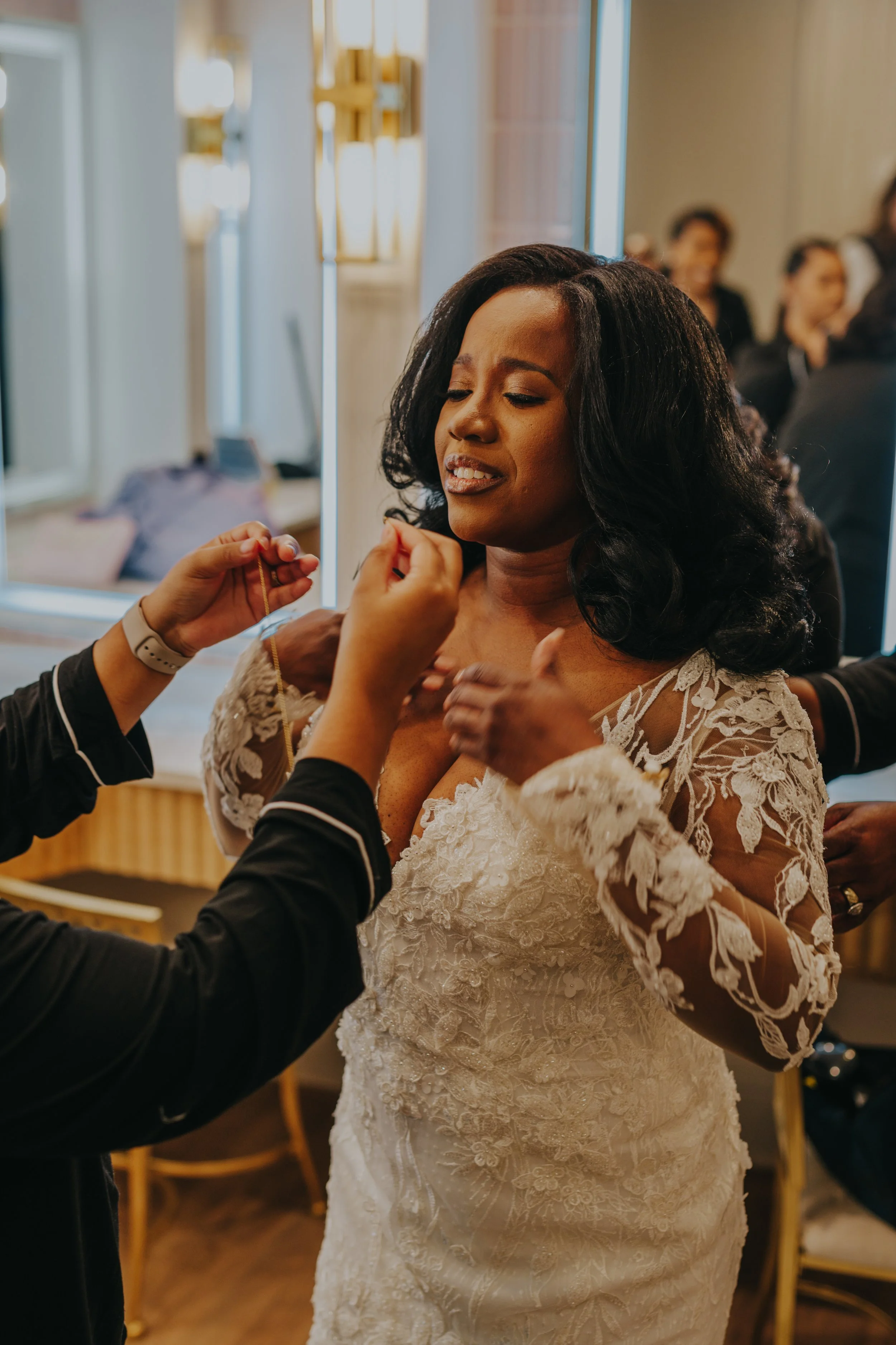 A woman in a white lace wedding dress is having her necklace put on by a person whose hands are visible. The woman has a satisfied expression. Other people are in the background, smiling and engaged in conversation.