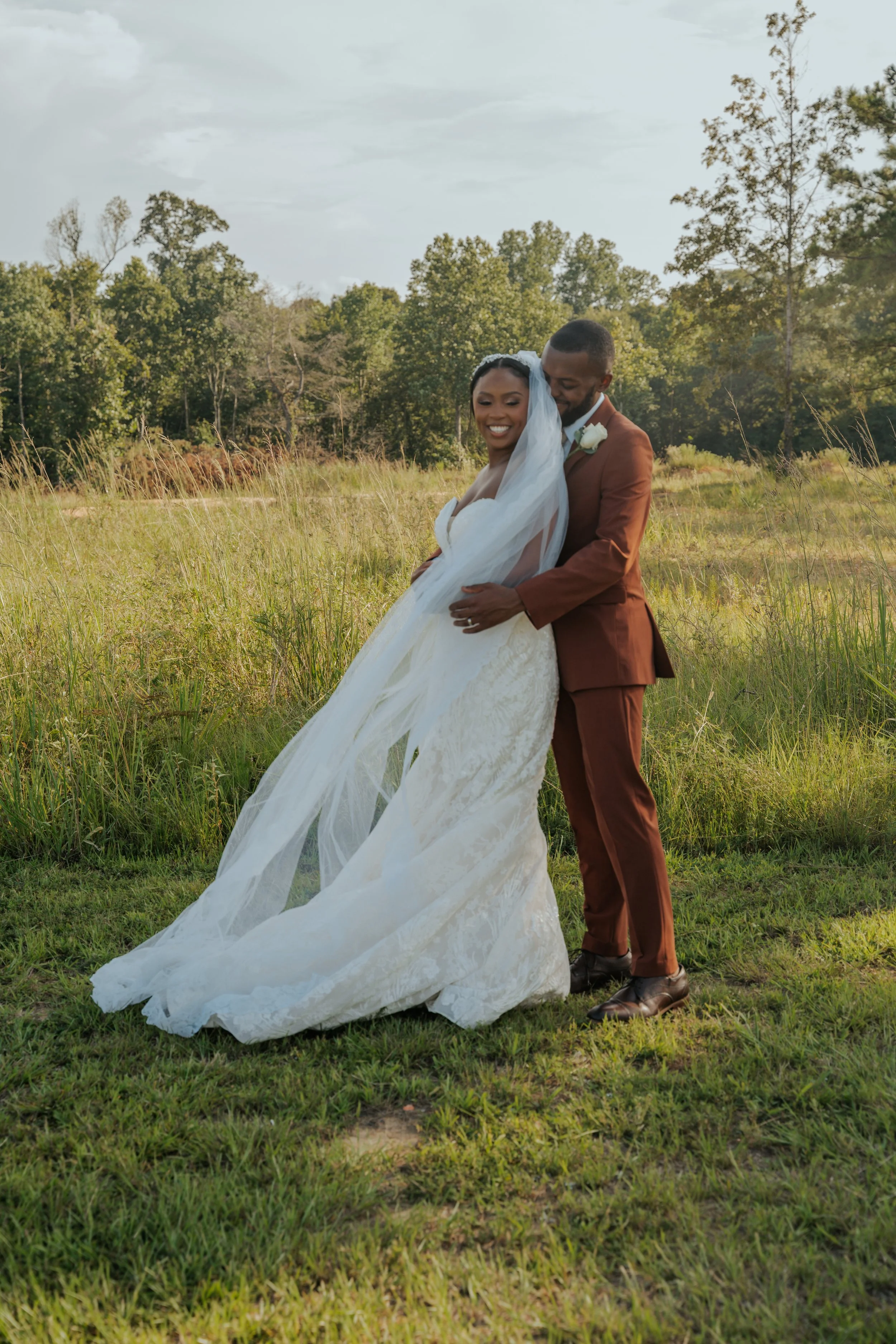 A couple dressed in wedding attire smiling and embracing outdoors in a grassy field with trees in the background.
