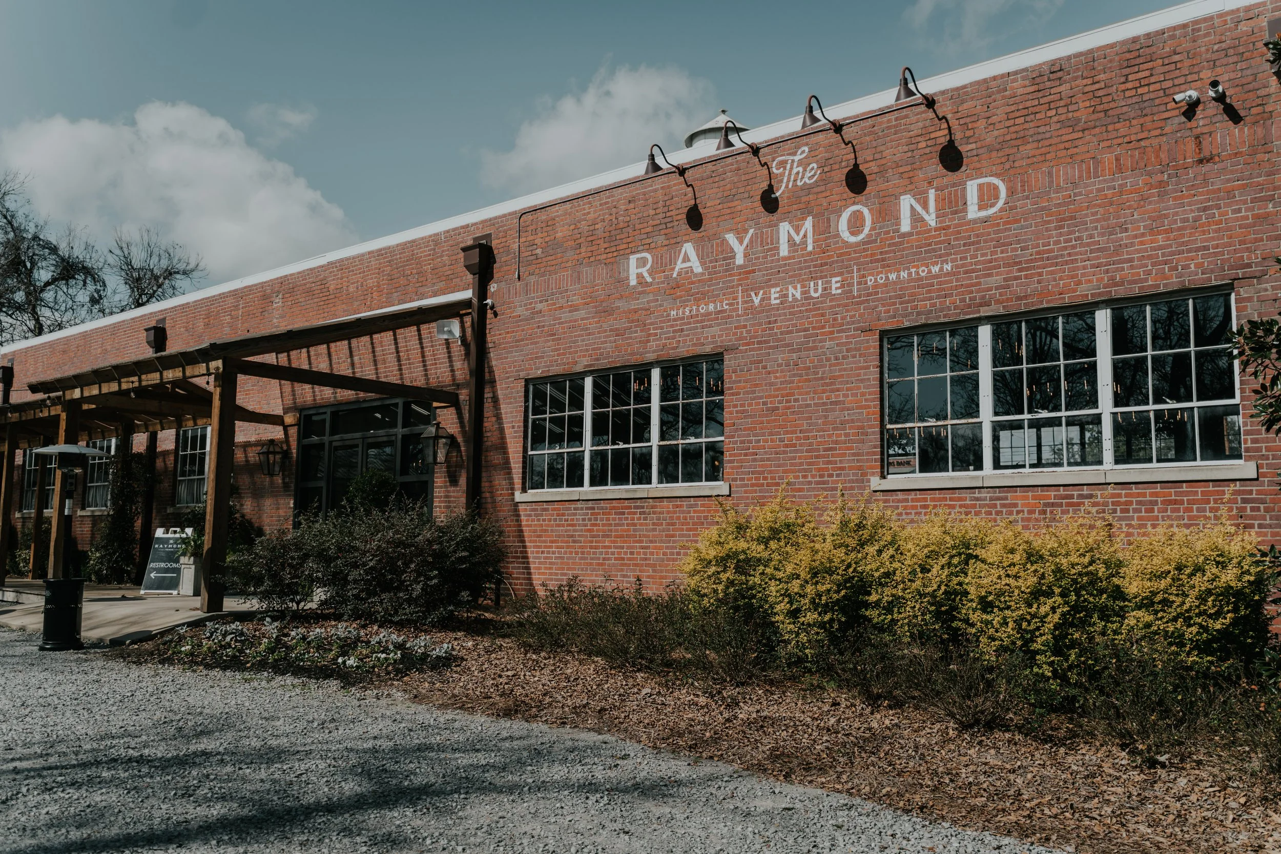 Exterior view of a brick building with the sign 'The Raymond' and smaller text 'Historic Venue Downtown'. A gravel pathway leads to the entrance, which has a covered porch with wooden beams and lantern-style lights. Bushes and plants are in front of 
