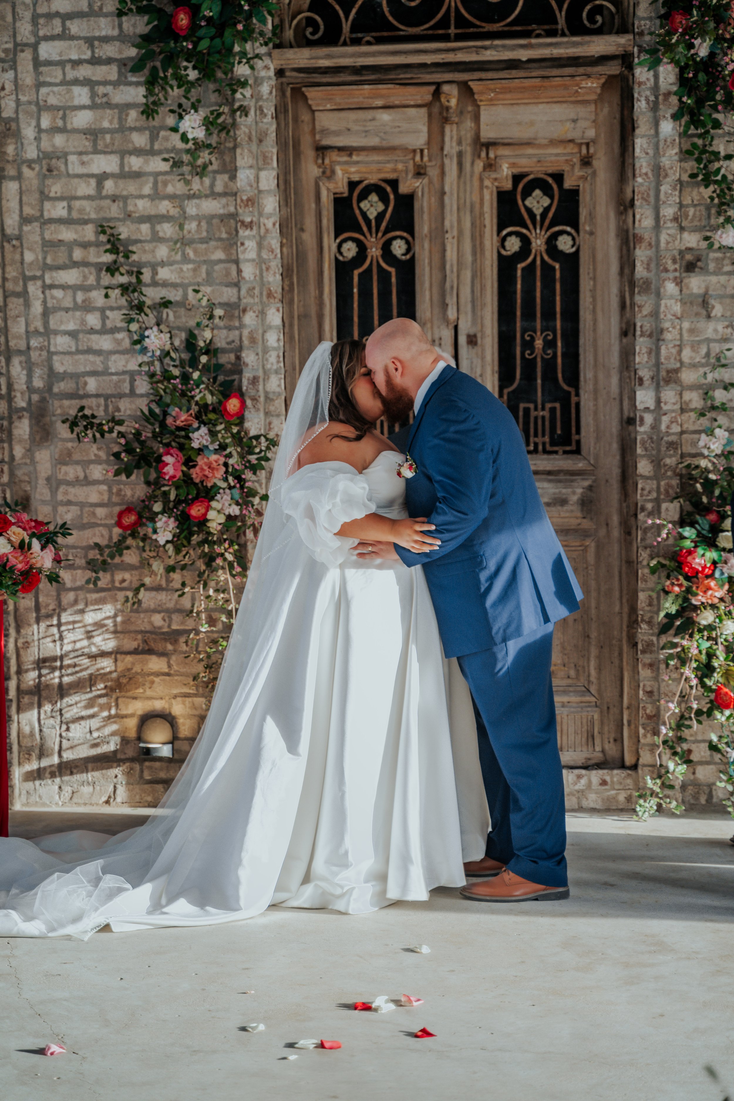 A bride and groom sharing a kiss during their wedding ceremony, standing in front of a rustic wooden door decorated with flowers and greenery, with flower petals scattered on the floor.