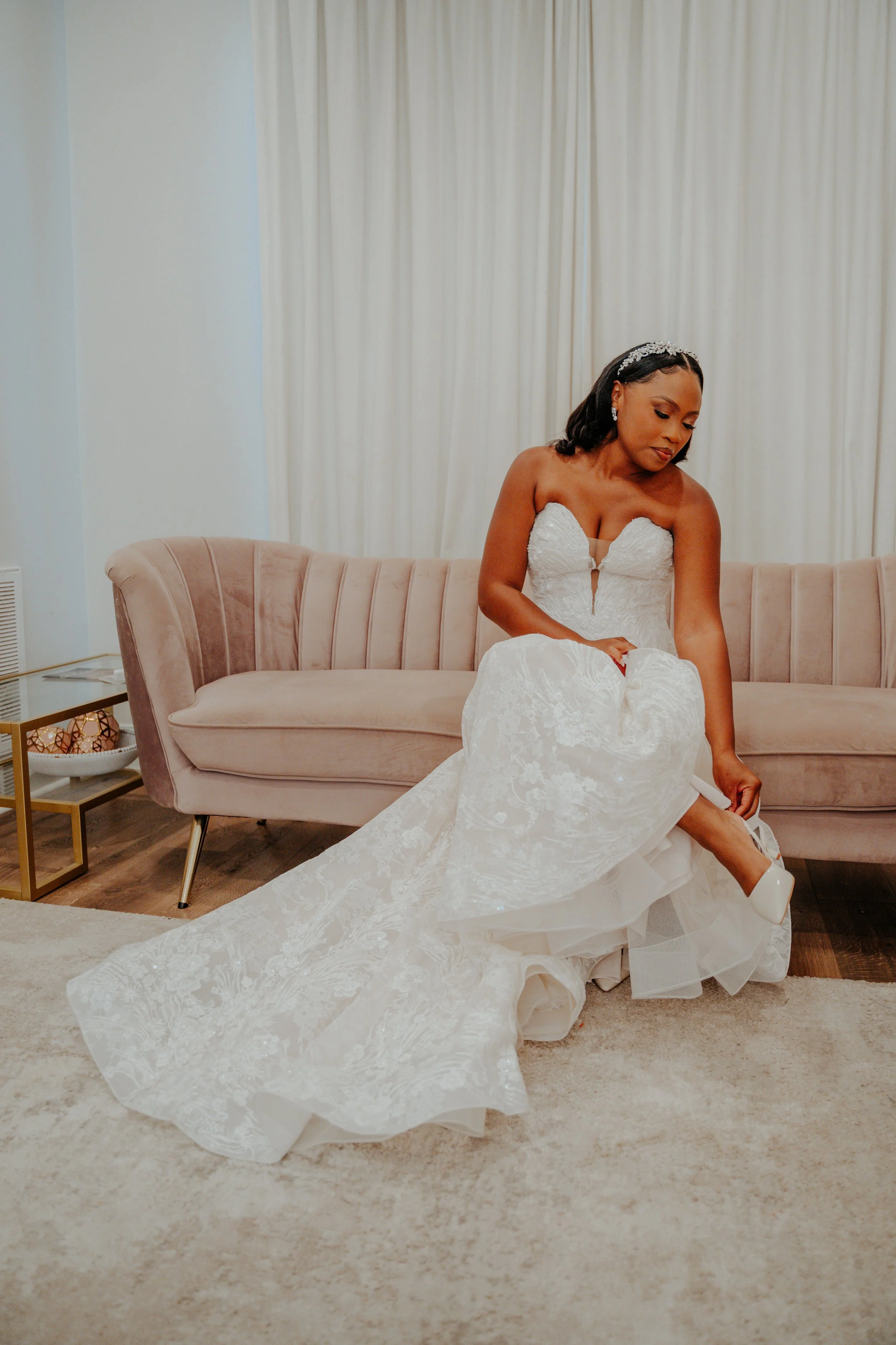 Bridal bride in a white wedding gown sitting on a pink velvet couch, adjusting her shoe.