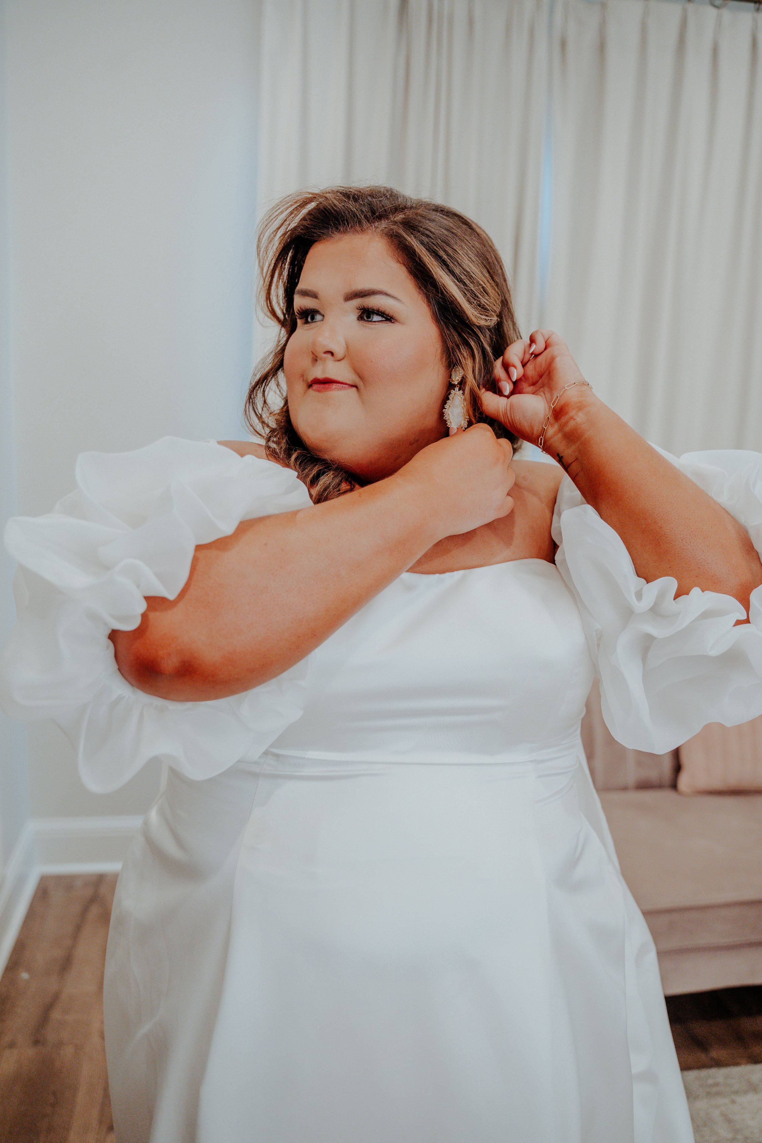 A woman in a white wedding dress putting on earrings.