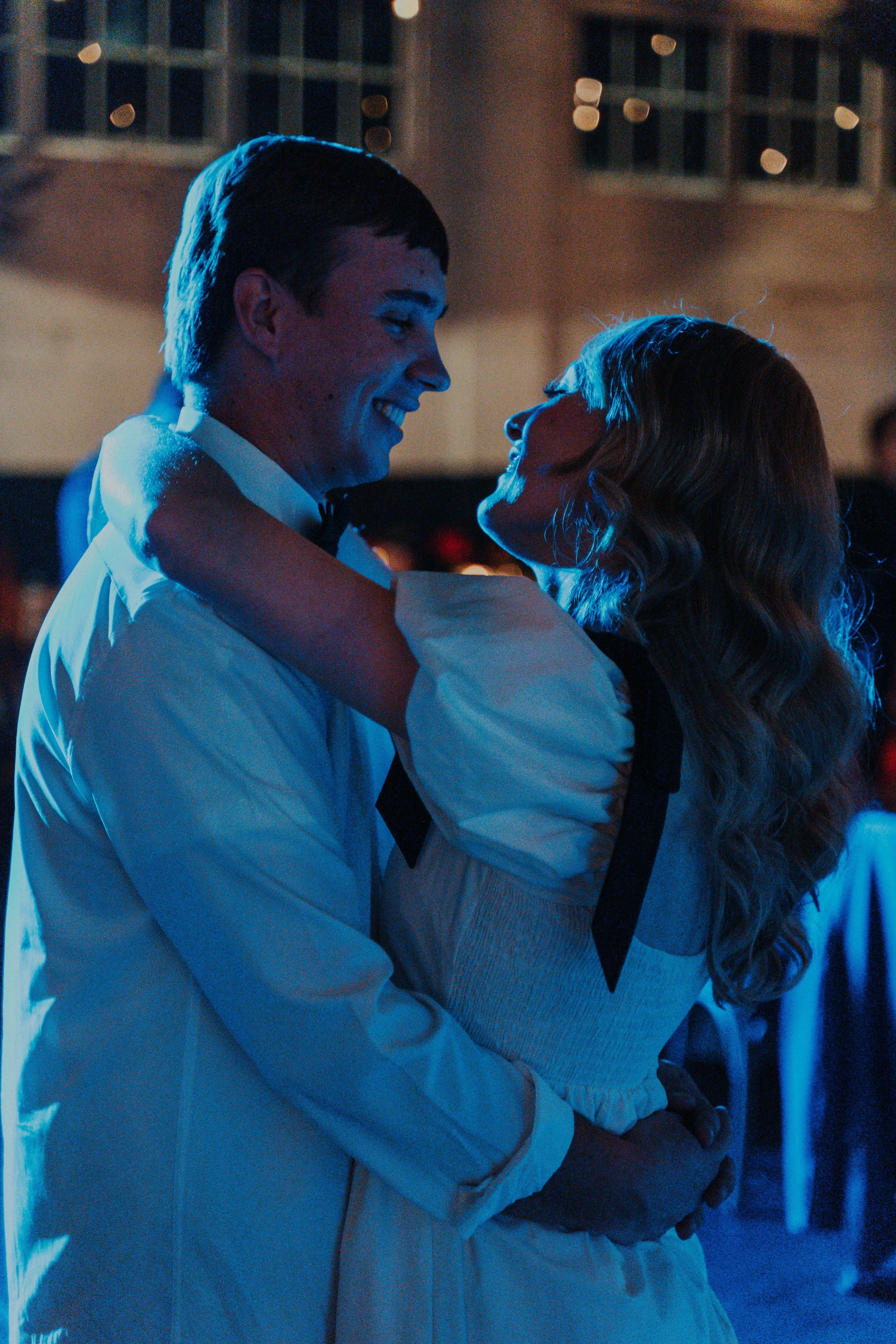 A young man and woman dance closely, smiling at each other, in a dimly lit setting with blue lighting and blurred background.