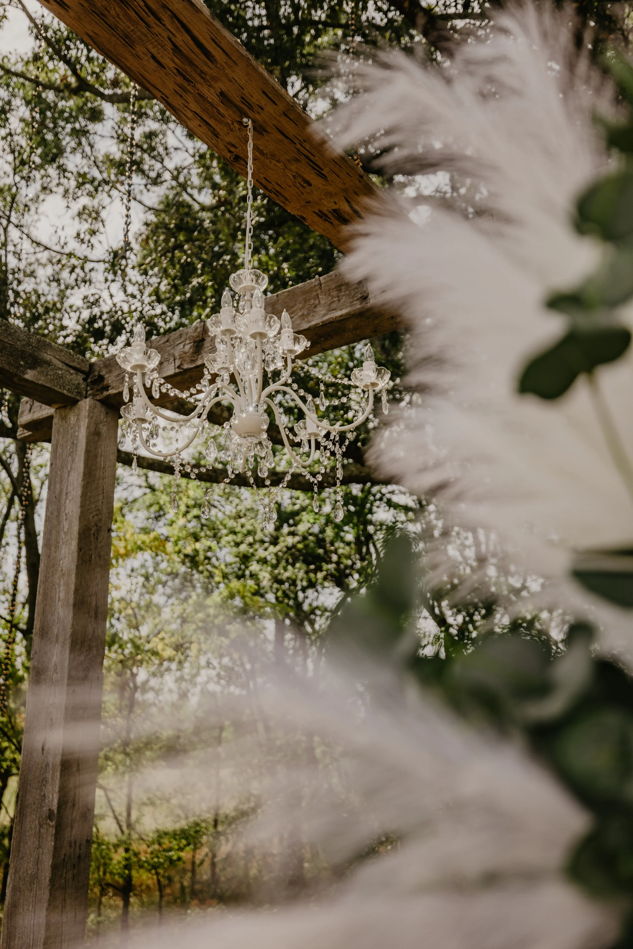 A white chandelier hanging from a wooden beam outdoors, with greenery and trees in the background, and some blurred white flowers and leaves in the foreground.