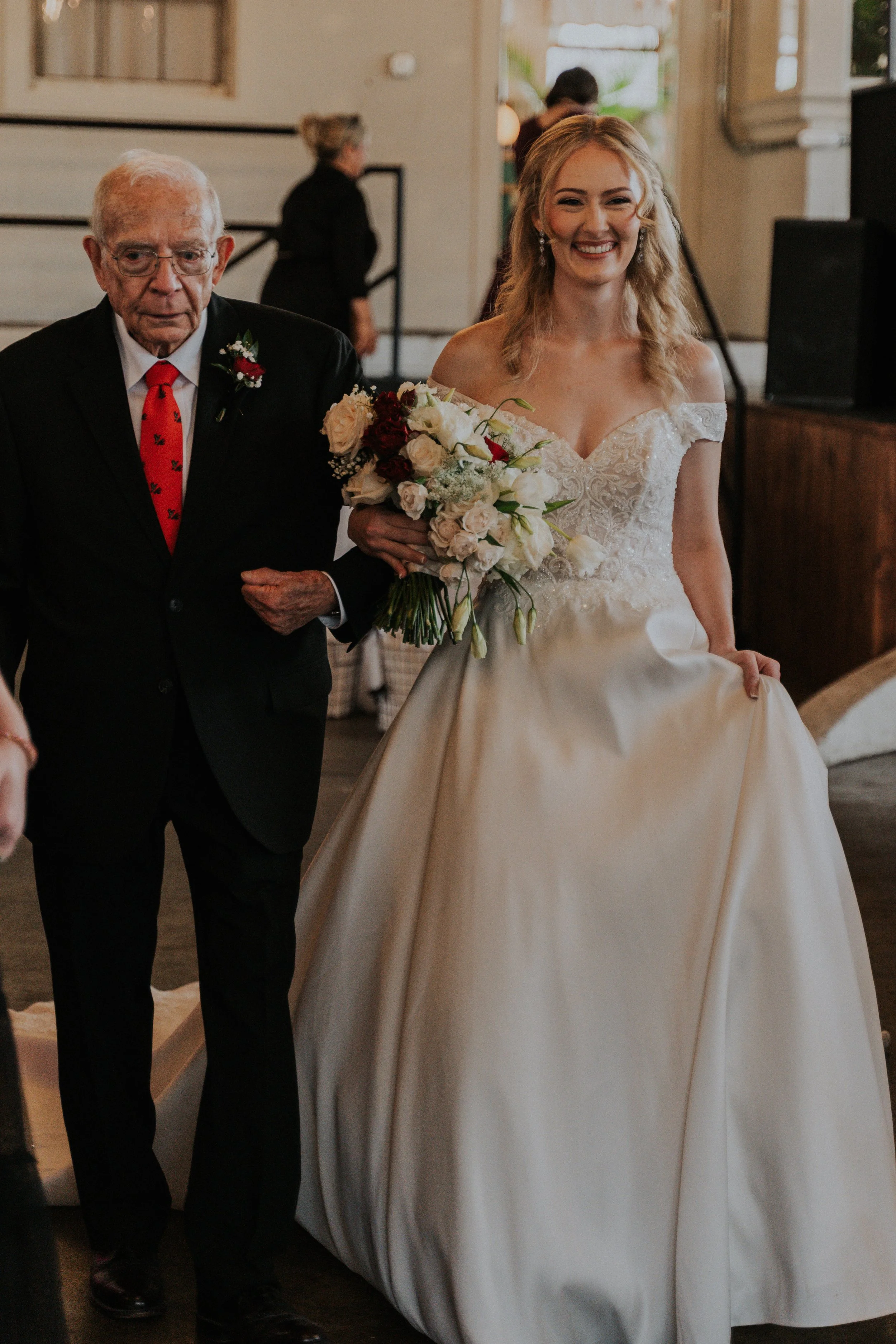 A bride in a white wedding dress smiling and holding a bouquet of flowers, walking down the aisle with an elderly man in a black suit and red tie.