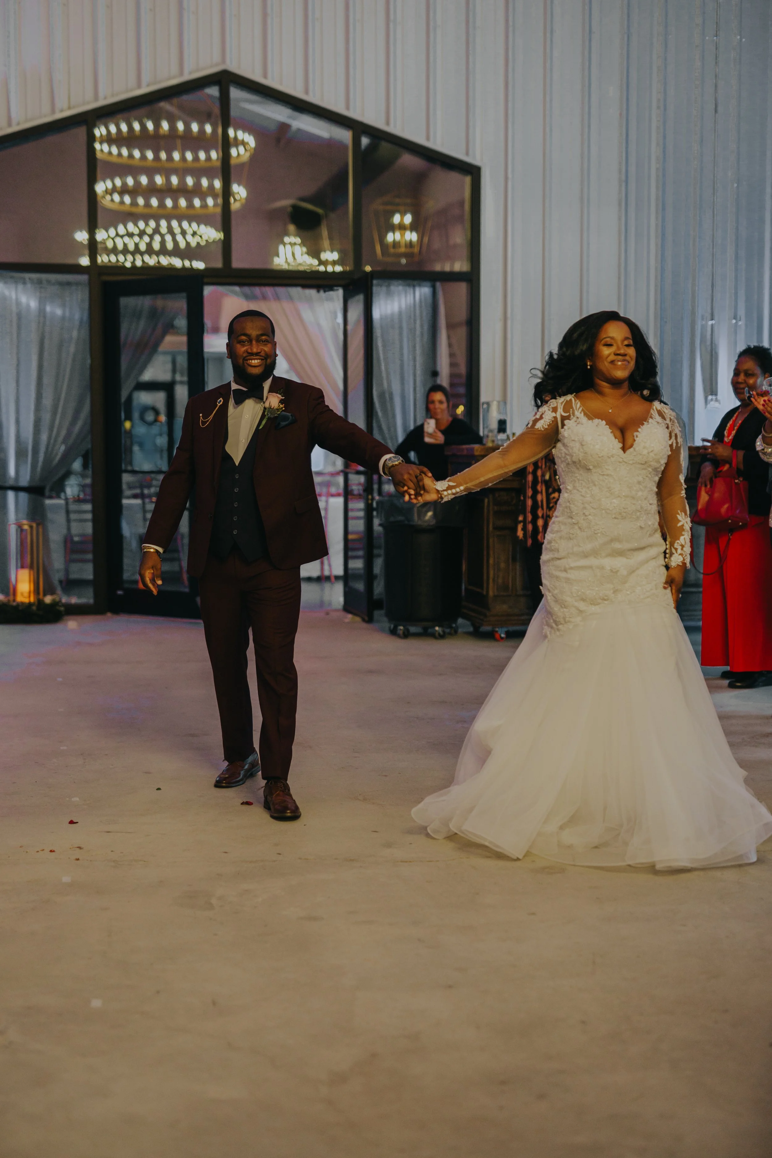 Bride and groom dancing at a wedding reception, holding hands, smiling, in a decorated indoor venue with guests watching.