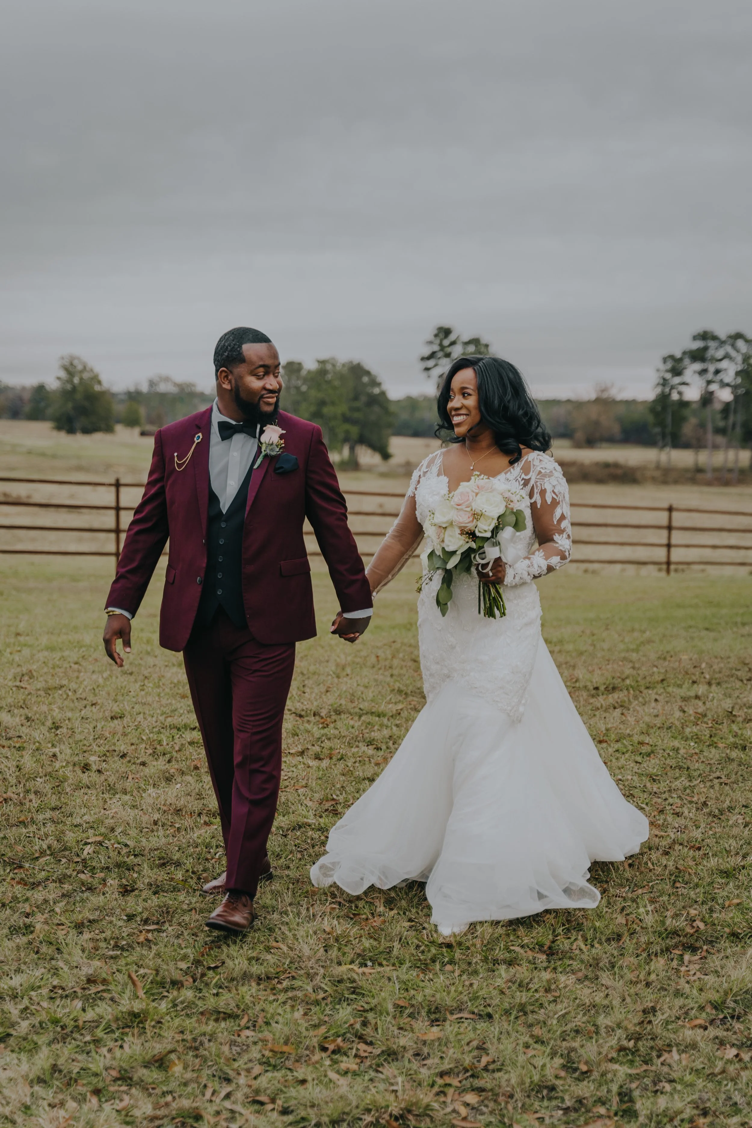 A bride and groom holding hands and smiling outdoors on a cloudy day. The bride is wearing a white wedding gown with lace details and holding a bouquet of flowers. The groom is dressed in a maroon tuxedo with a black bow tie, and they are walking acr