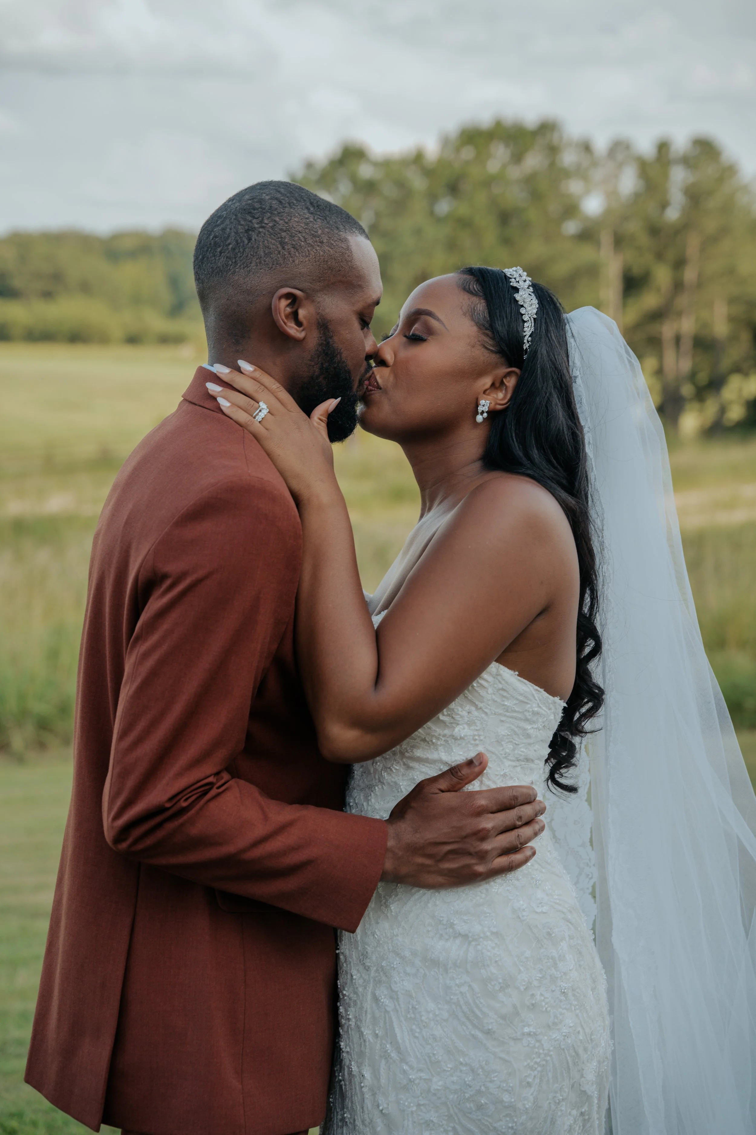 A bride and groom sharing a kiss outdoors in a natural setting, with the bride wearing a white strapless wedding gown, veil, and jewelry, and the groom in a rust-colored suit.