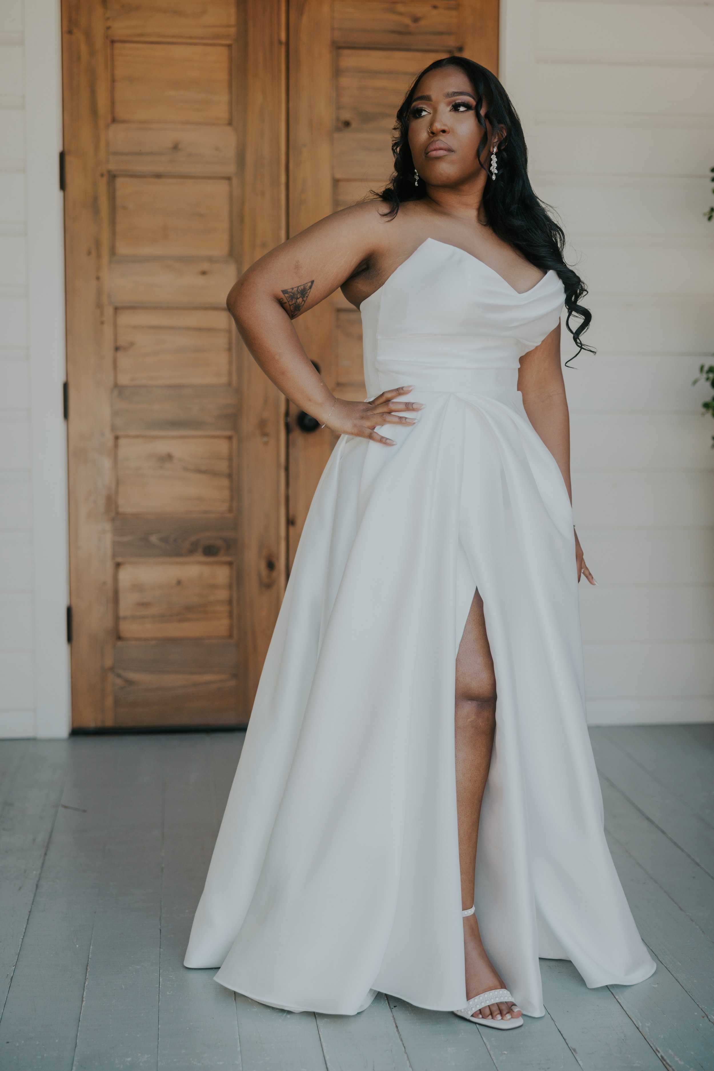 A woman in a white wedding dress standing indoors with one hand on her hip and looking to the side. She has dark hair, earrings, and a tattoo on her upper left arm. The background features a wooden door and white wall paneling.