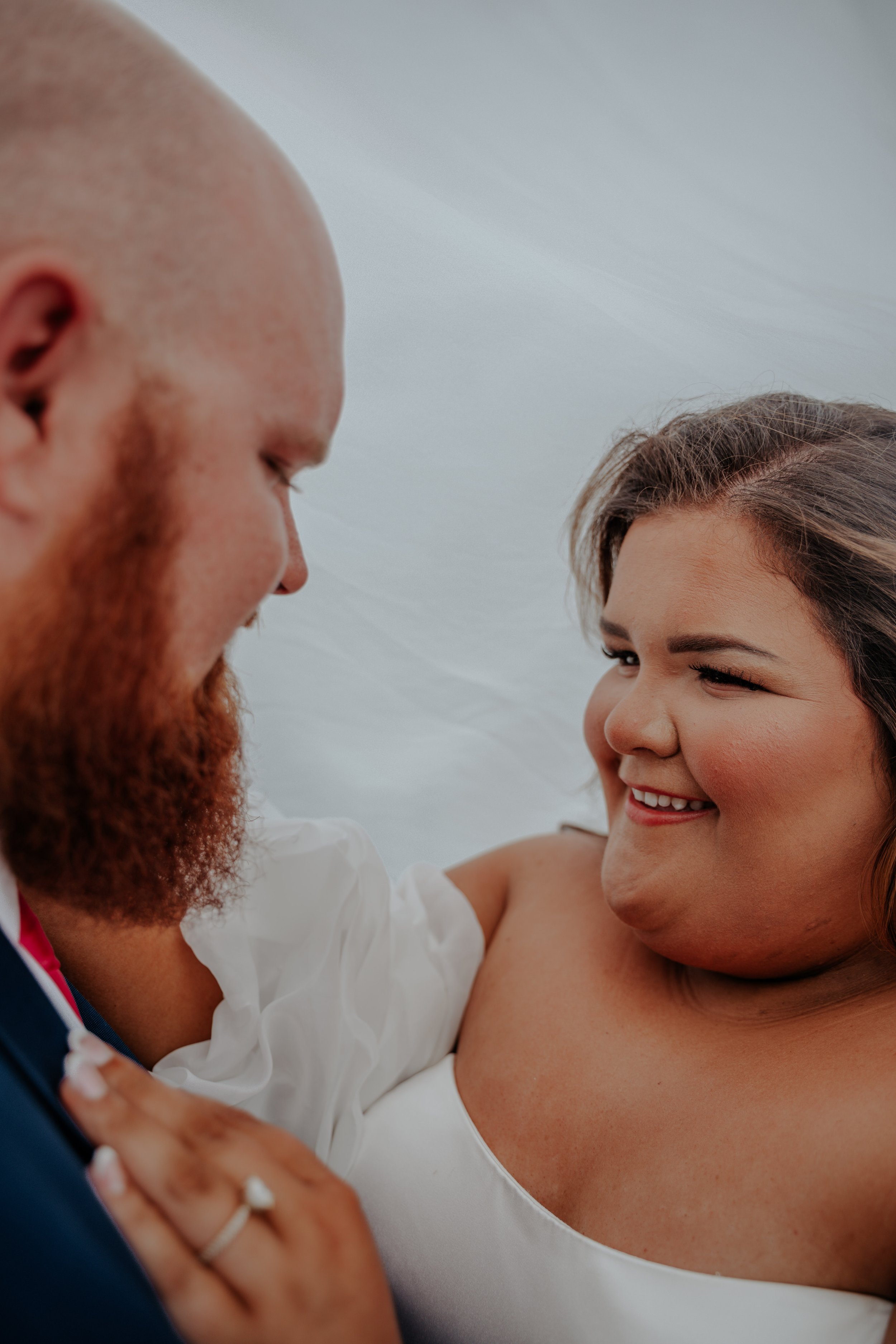 A couple on their wedding day, smiling and looking at each other closely, with the bride wearing a white dress and the groom in a suit.